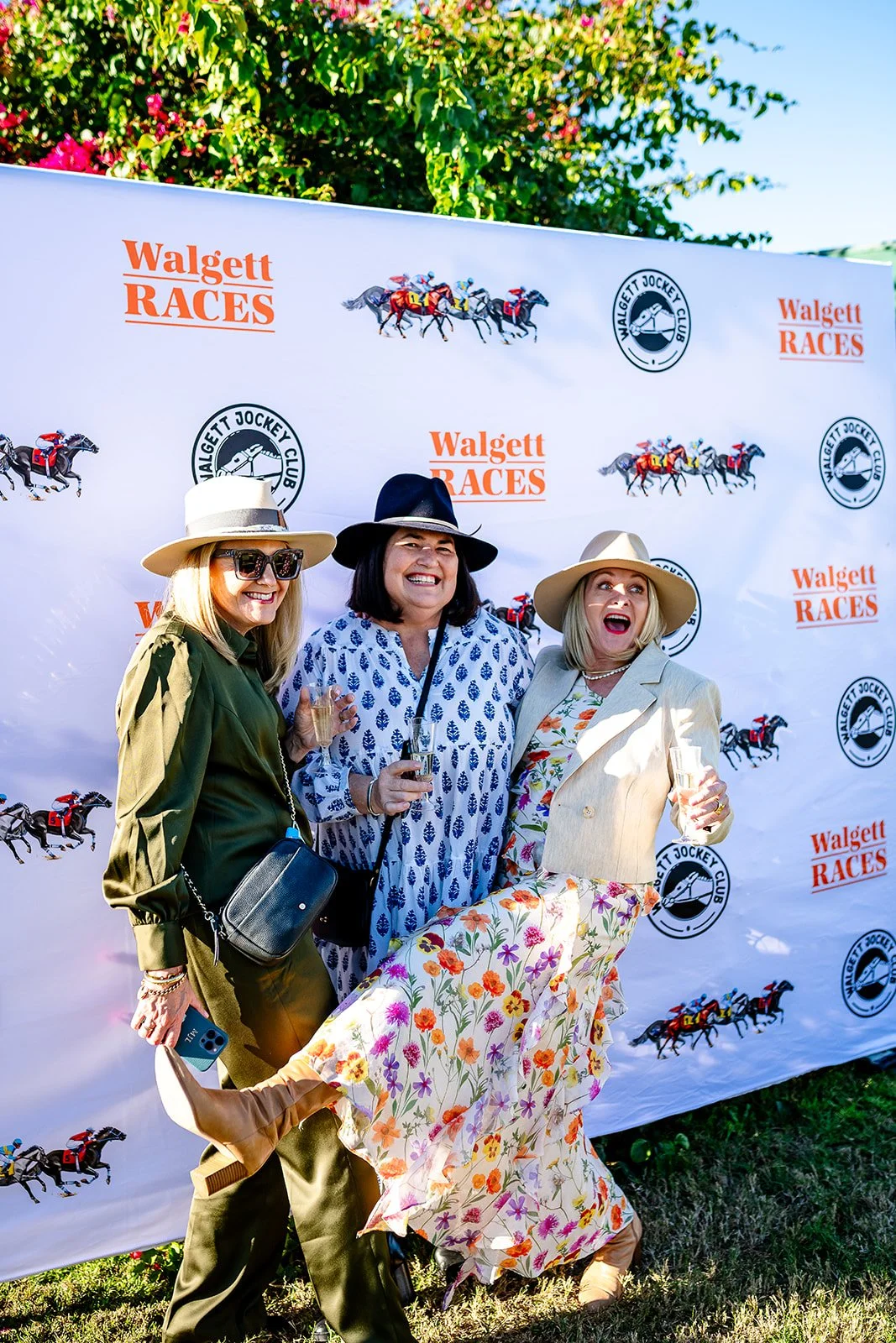 Fashions destination at the Walgett Races | Three women dressed in fashionable attire and wearing hats, posing with drinks in front of a backdrop with horse racing logos, at an outdoor event.