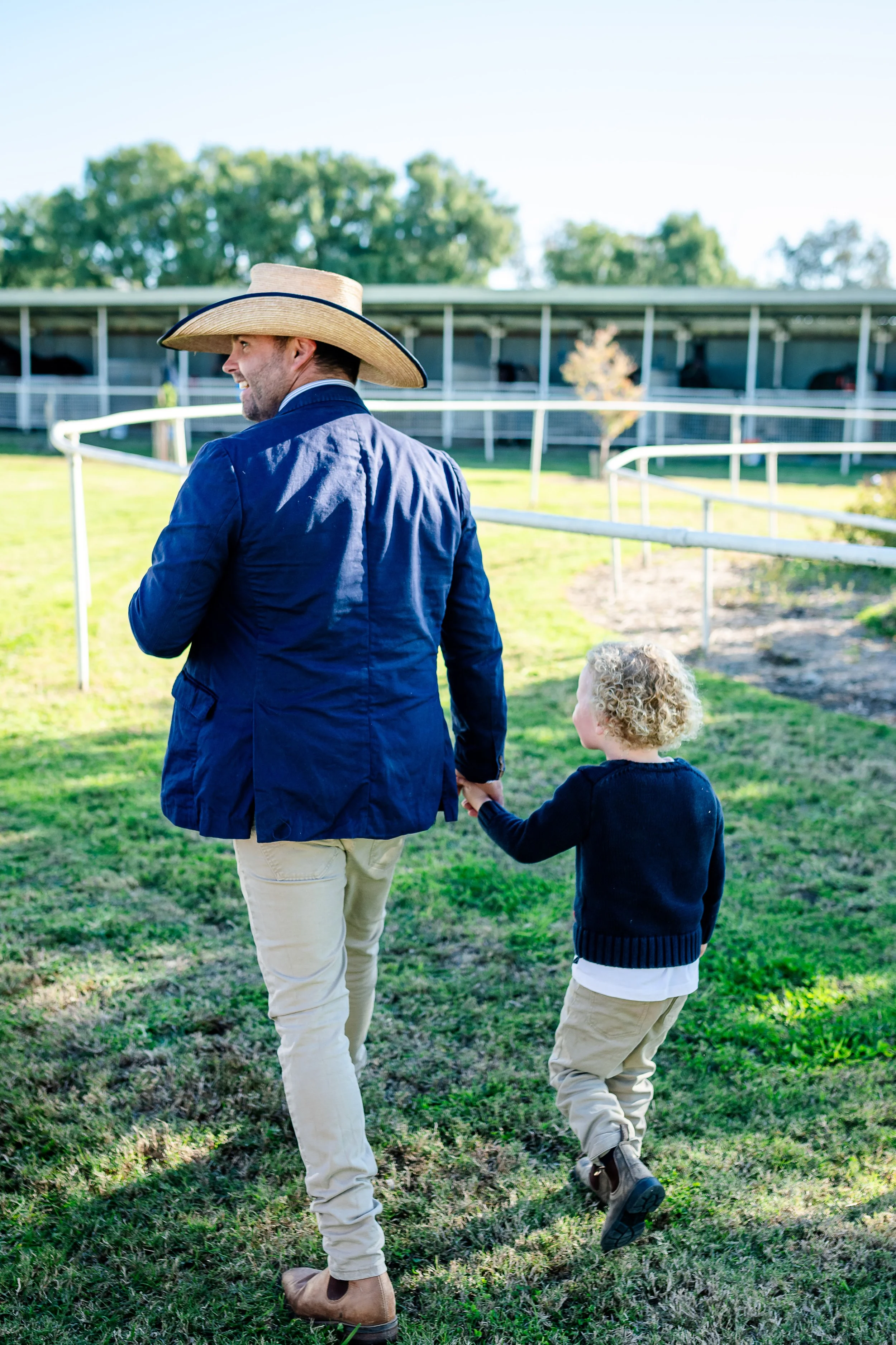 A man with a large sun hat holding the hand of a young boy at the Walgett Races, walking on grass near a horse stable on a sunny day.