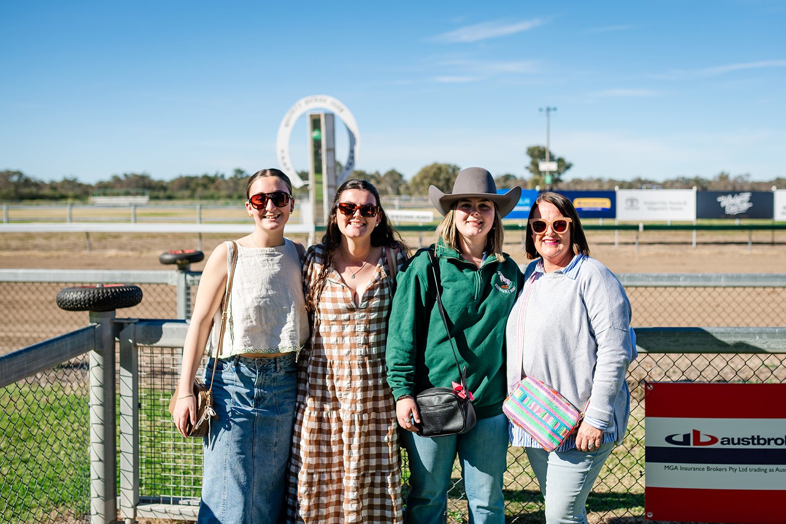 Four women standing side by side outdoors at the Walgett Races racetrack on a sunny day, smiling at the camera, with a fence and the racetrack in the background.