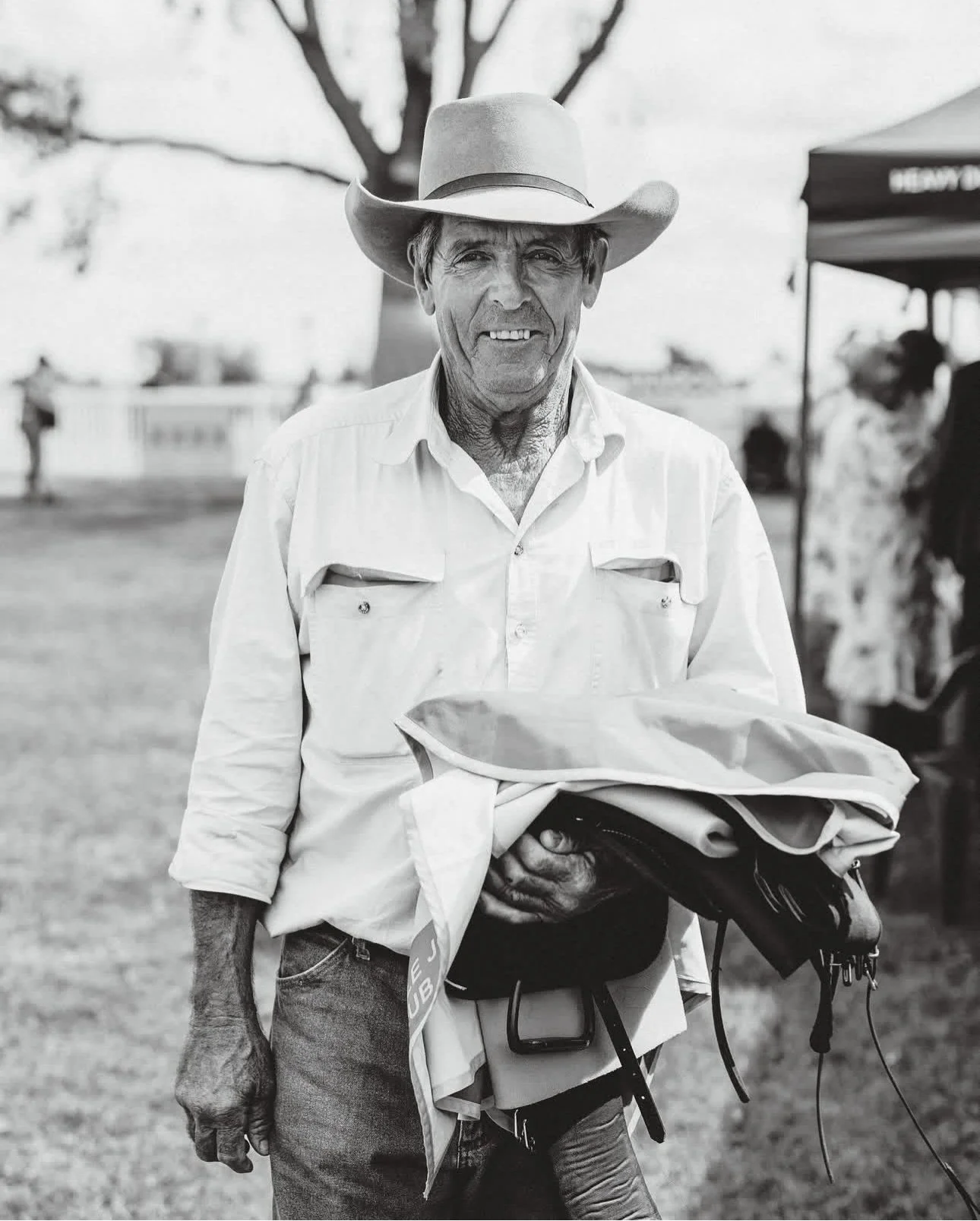Kelly Smith wearing a cowboy hat and a light-colored button-up shirt stands outdoors at the Walgett Races, holding folded clothing and other items in his hands, with trees and a tent in the background.
