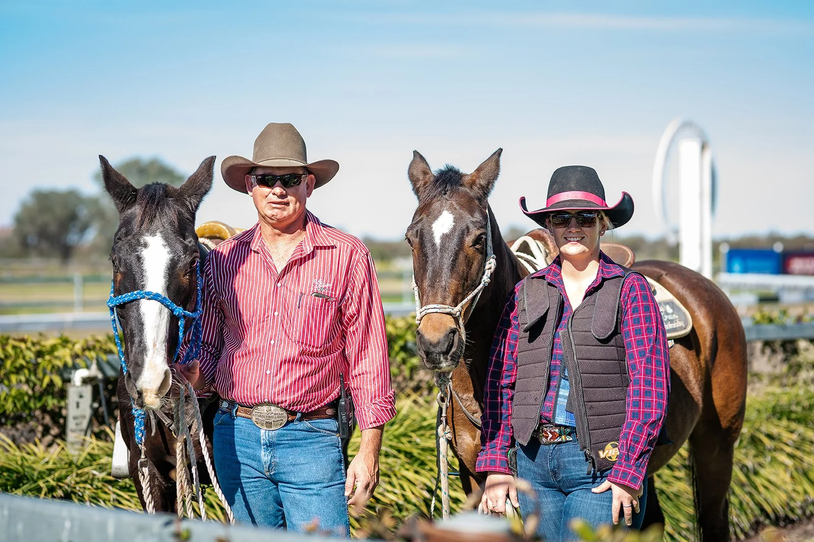Two people, a man and a woman, stand next to horses the Walgett Races. The man wears a wide-brimmed hat, sunglasses, a red striped shirt, and jeans, while the woman wears a black hat with a pink ribbon, sunglasses, a plaid shirt, and a vest.