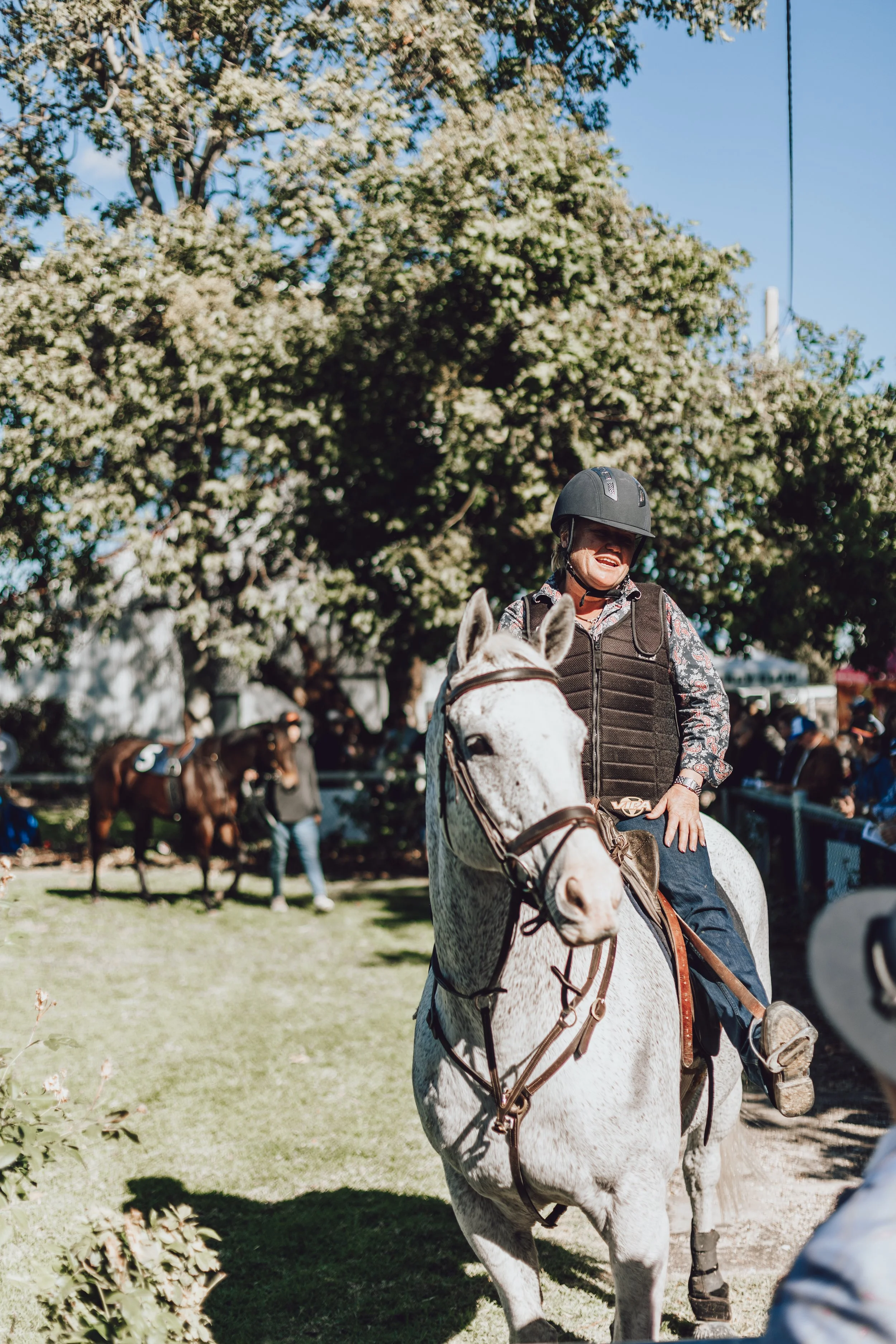 Mel Ozoux wearing a black helmet and vest riding a white horse on a sunny day, with other horses and people in the background, at the Walgett Races