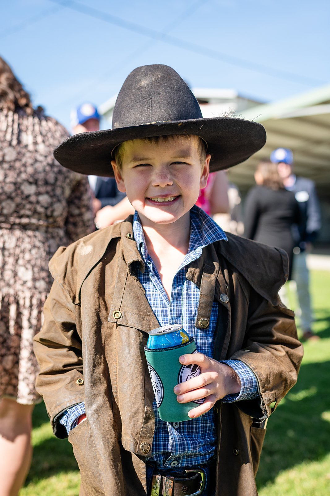Young boy dressed in cowboy attire, wearing a large black cowboy hat, plaid shirt, and brown jacket, smiling with a drink can in a green koozie in hand at an outdoor event on a sunny day.