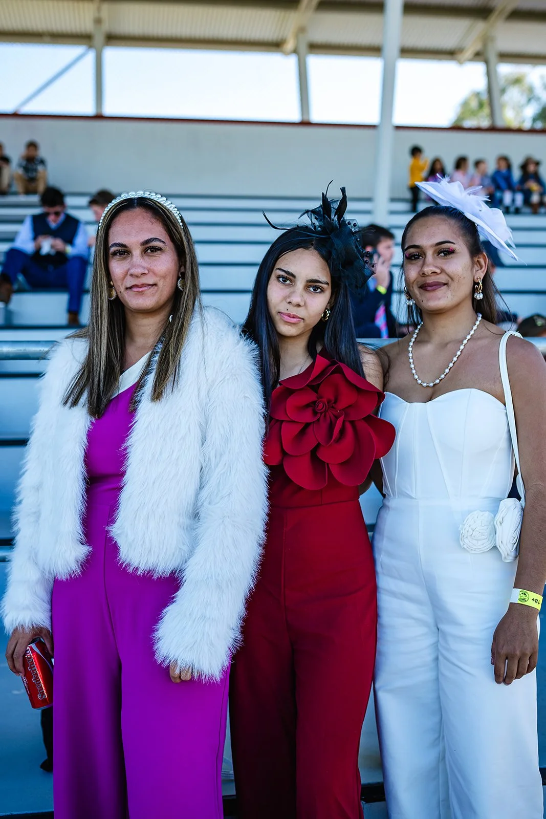Three women dressed in formal attire standing together at the Walgett Races with stadium seating in the background.