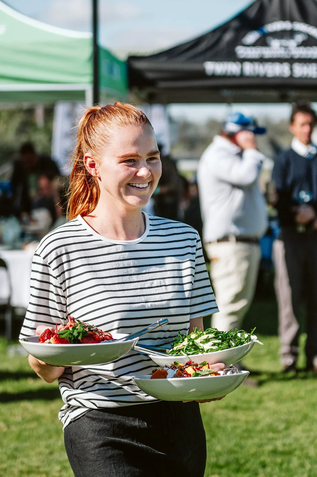 A woman smiling and carrying three bowls of salad at the ticket luncheon at the Walgett Races, with tents and other people in the background.