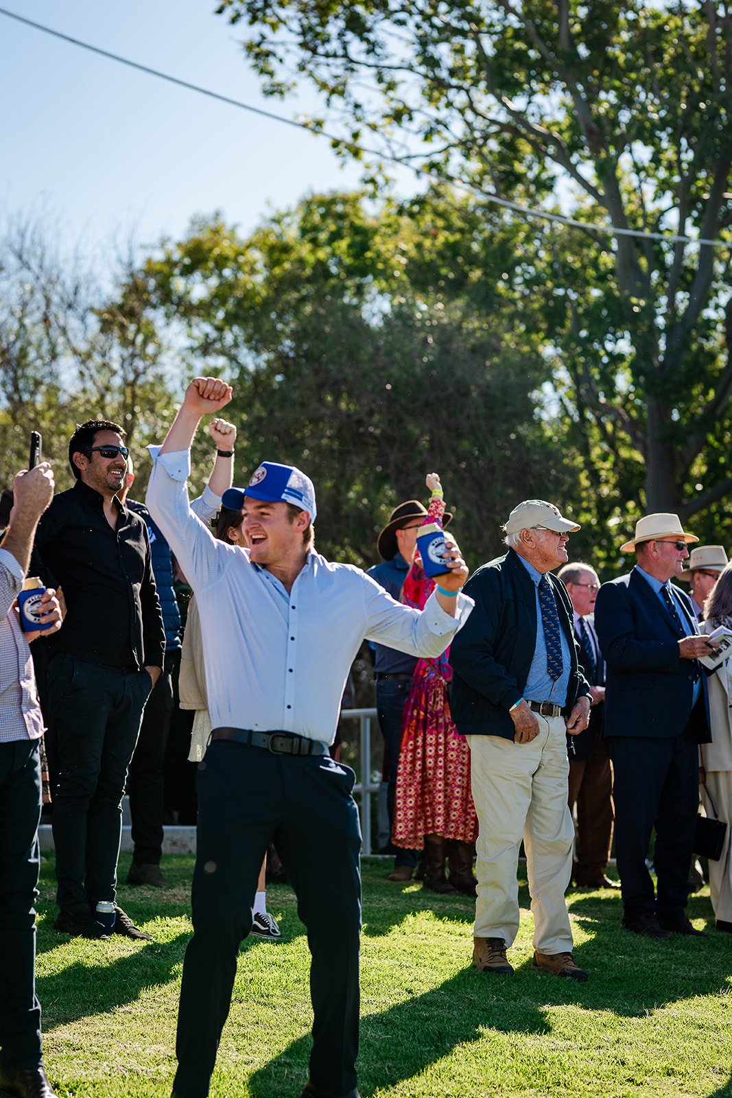 Celebration of a young man in a white shirt with a blue cap holding a drink, cheering with others at the Walgett Races on a sunny day with trees in the background.