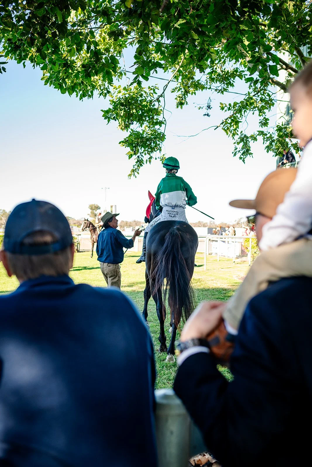 A jockey from the  Walgett Races dressed in green and white riding gear on a horse at a racetrack, with several spectators watching under a leafy tree on a sunny day.
