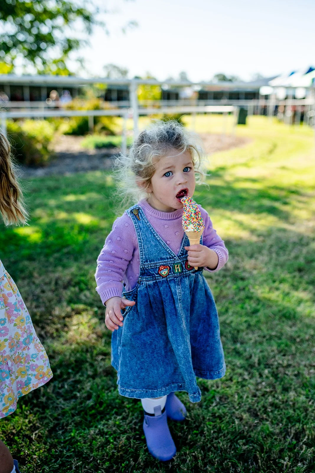 A young girl with messy blonde hair and blue eyes is standing on grass at the Walgett Races, holding and eating a colorful ice cream cone.