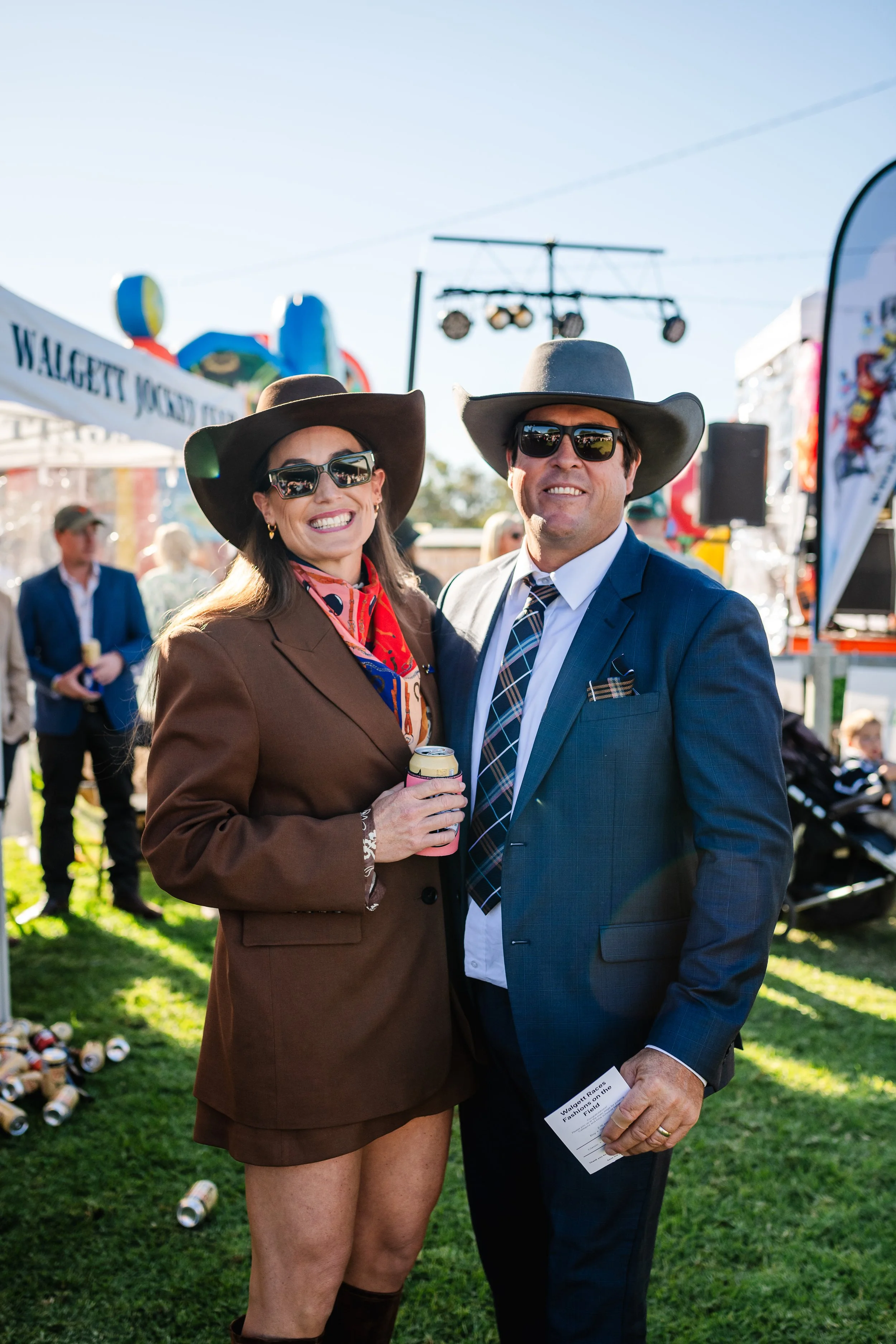 A smiling woman and man dressed in Western-style cowboy hats and formal attire, standing outdoors at an event. The woman is holding a beverage can, and the man is holding event tickets, with other people and event tents visible in the background.