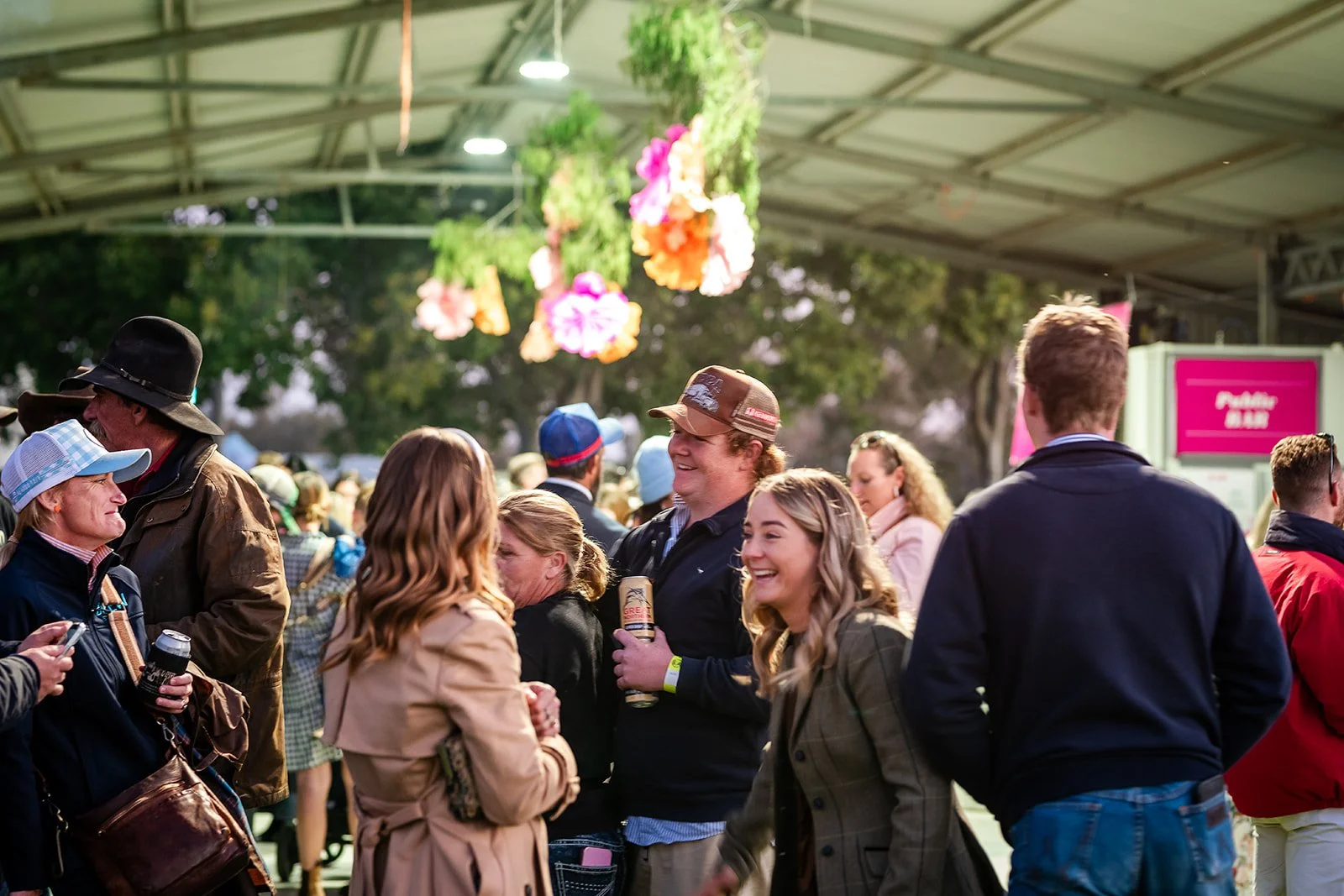 People socializing at an outdoor festival under a tent with hanging flower decorations, some holding drinks, smiling and talking.
