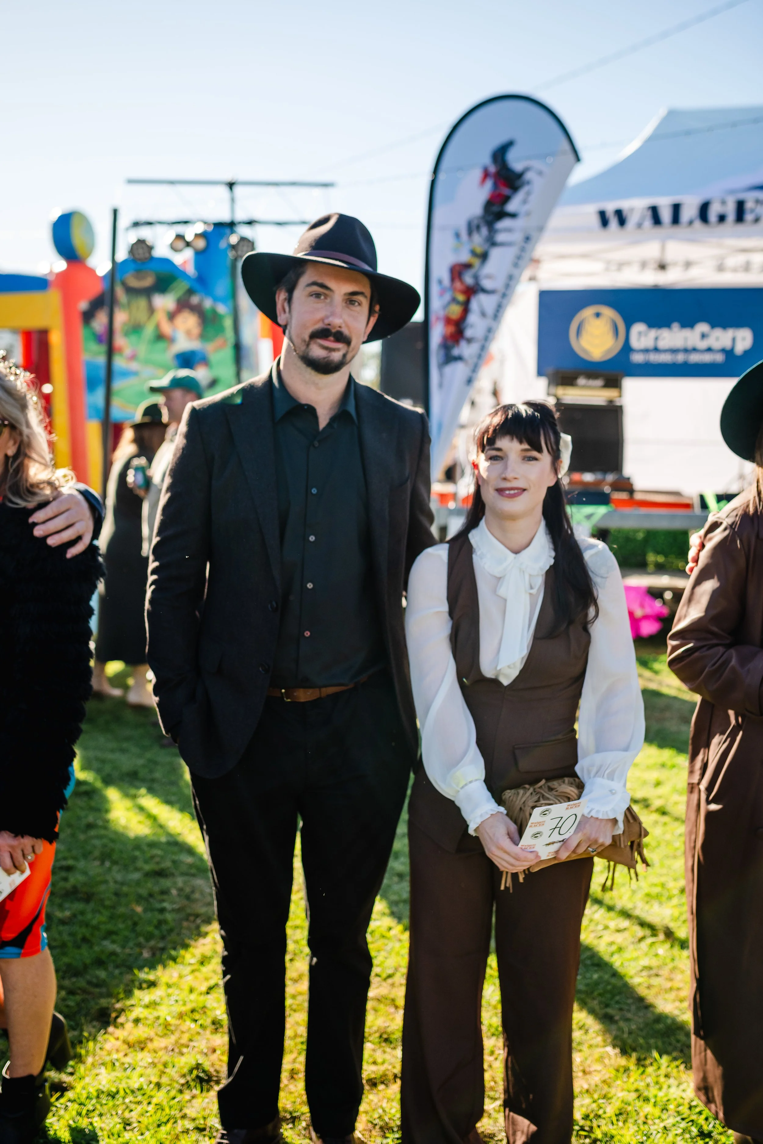 A man and a woman dressed in vintage-style clothing standing outdoors at the Walgett Races with game booths and flags in the background.