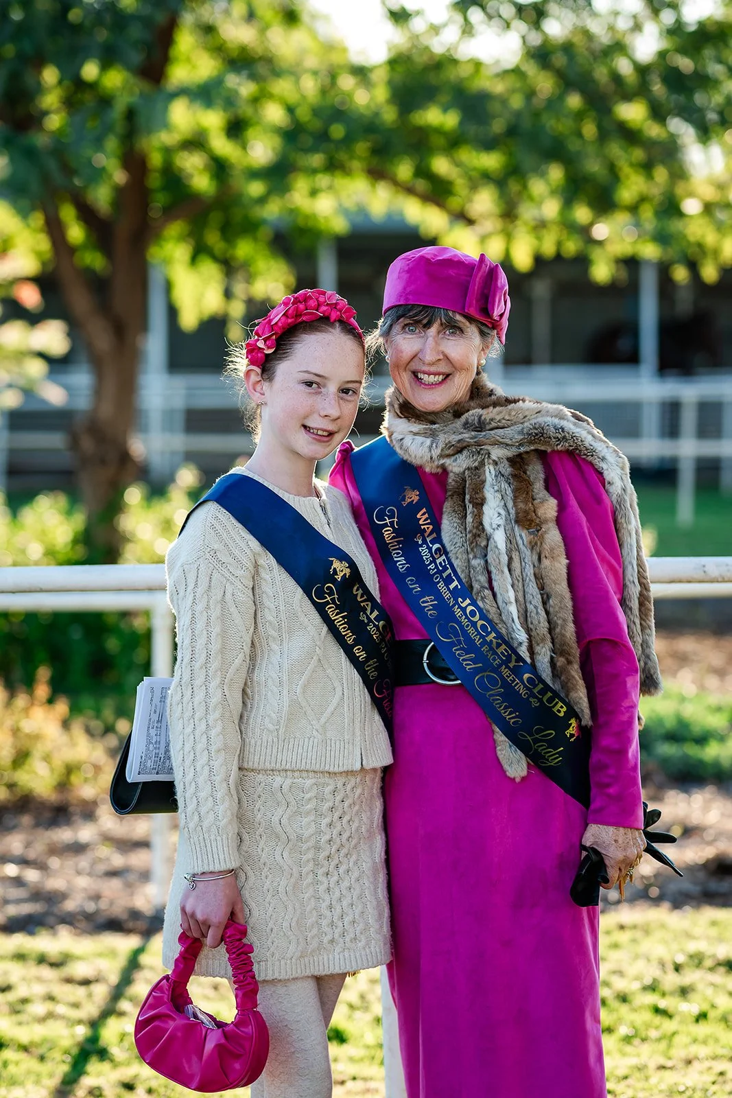 Two women, one younger and one older, standing side by side outdoors, smiling at the camera. They are dressed in vibrant pink and cream-colored outfits, wearing satin sashes that read 'WALGET JOCKEY CLUB' and 'Fashions on the Field.' The younger girl