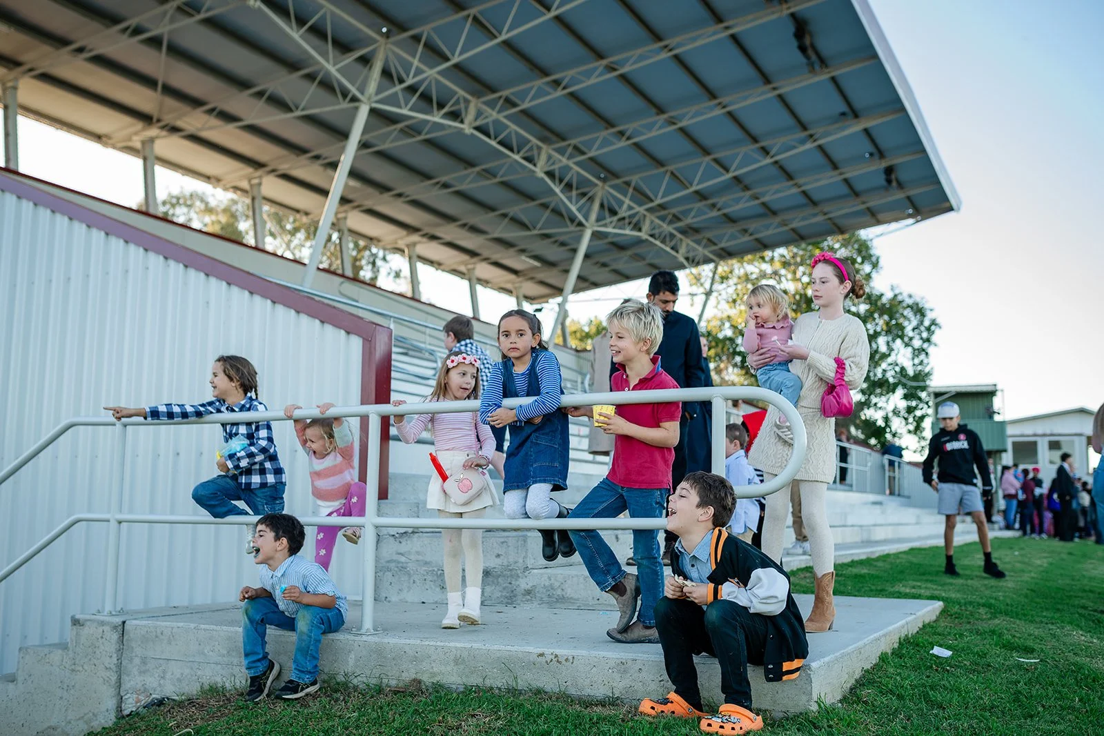 Children and adults standing and sitting on concrete stairs near a covered grandstand, with some children smiling and playing, during the Walgett Races.