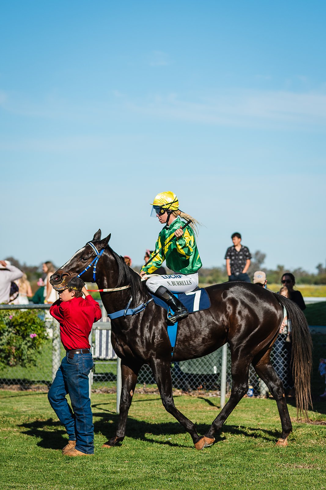 A jockey in green and yellow riding gear rides a dark brown horse at the Walgett Races, with a woman in red holding the horse's bridle and a small crowd watching in the background.