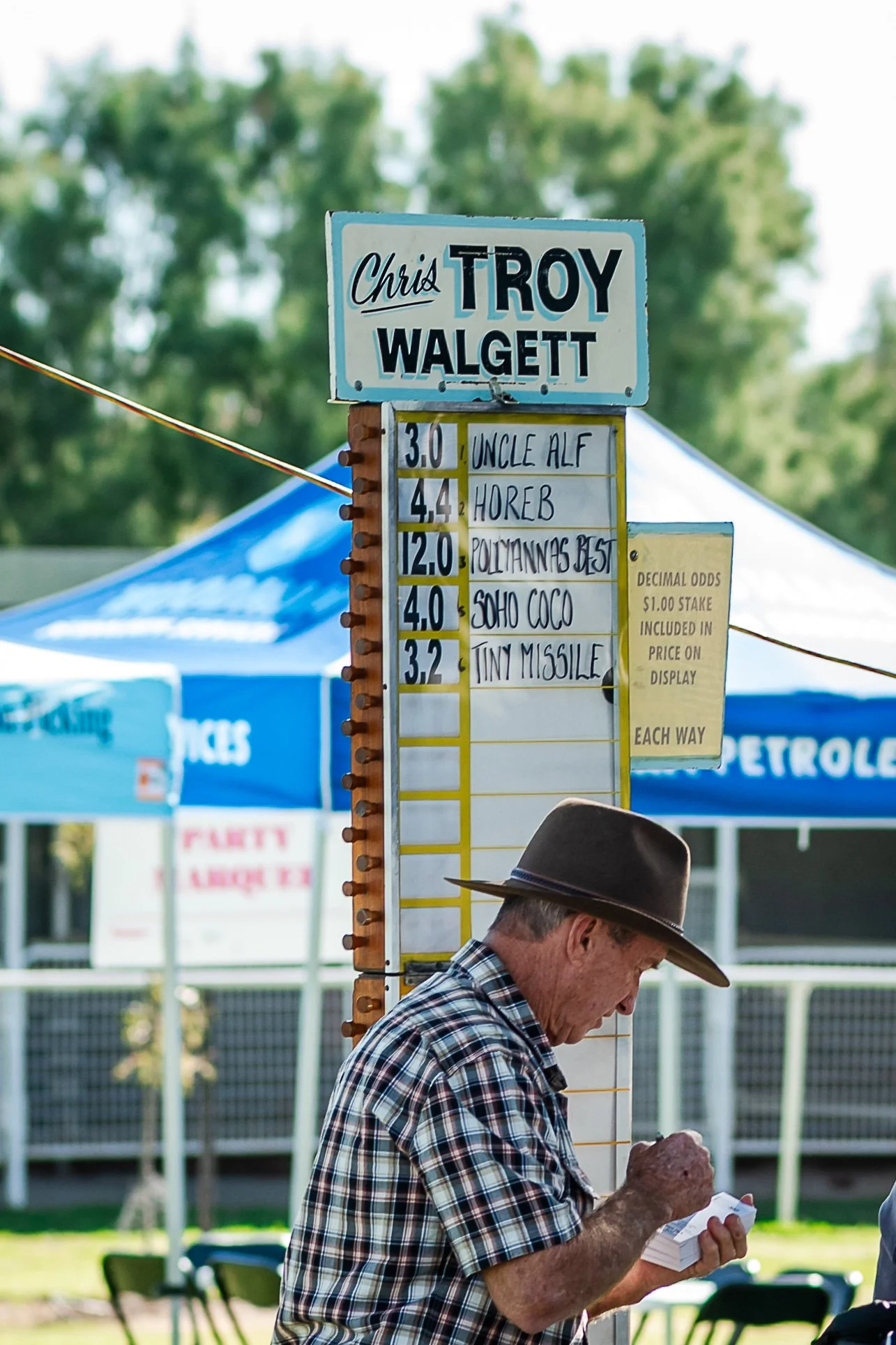 Chris Troy at the Walgett Races in a plaid shirt and wide-brimmed hat is looking at his booklet for his bookmaking. The scoreboard displays race horse names and their respective odds, with a background of blue tents and greenery.