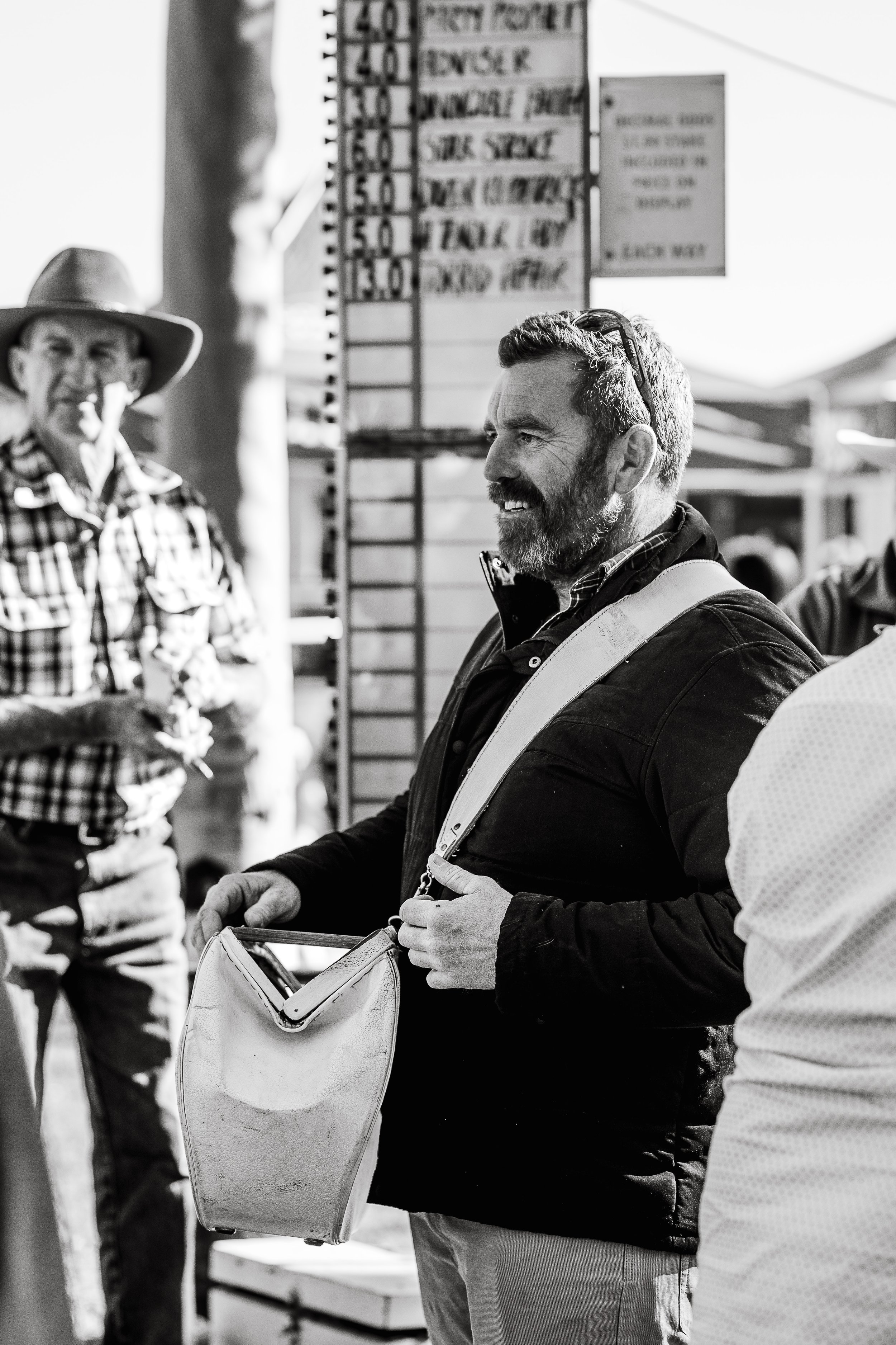 A man with a beard and short hair smiling at an outdoor market, holding a leather bag, with another man in a cowboy hat and checkered shirt in the background.