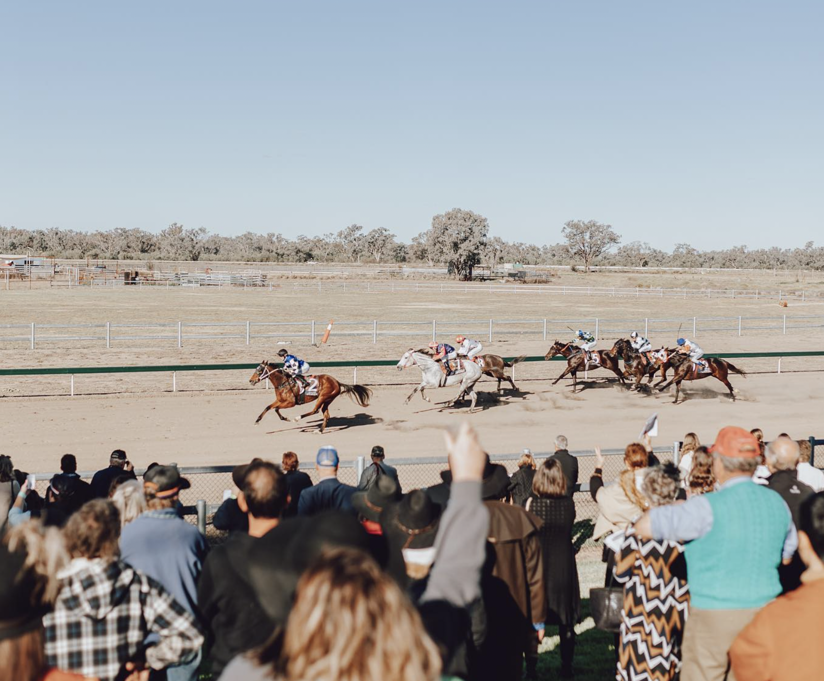Walgett Races | Horse racers competing in a race on a dirt track with spectators watching from behind a fence.