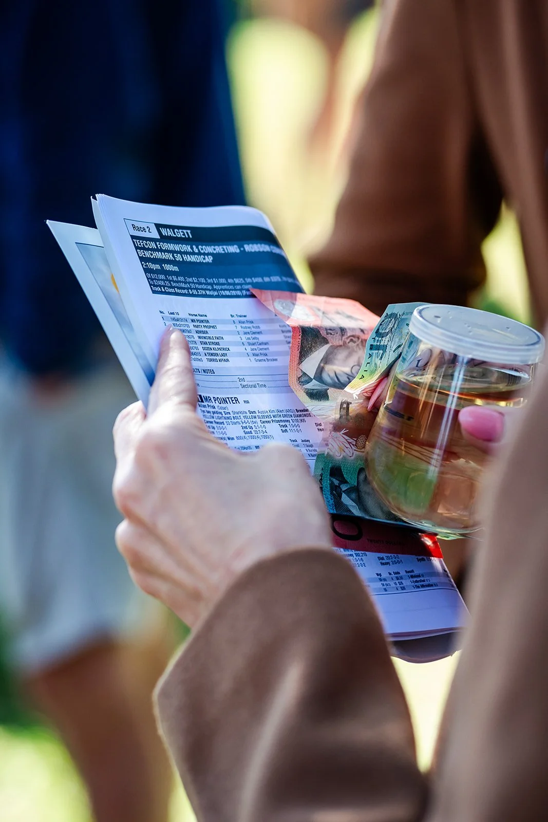 A person holding racing event tickets, a clear plastic cup with a drink, and a banknote. Country races at Walgett races. 