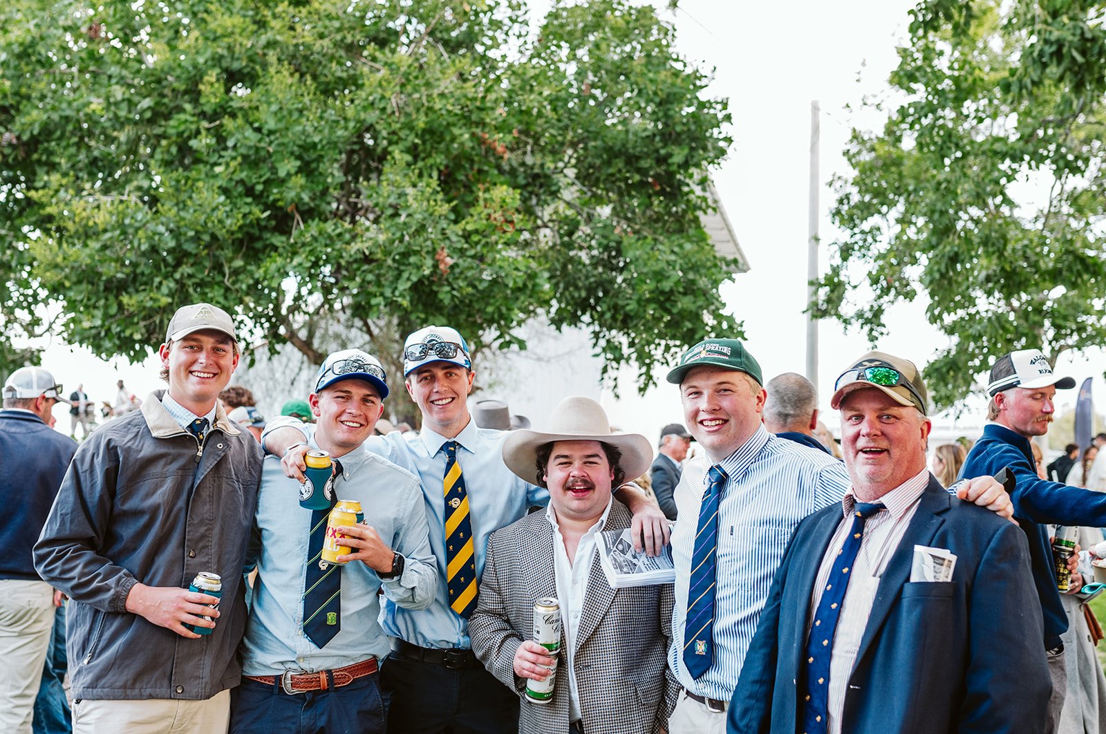 Group of seven men posing Walgett Races in Bill's Bunker, the best country race day in NSW. They are smiling, with drinks in hand, dressed in casual and semi-formal attire, under a large green tree with other people in the background.