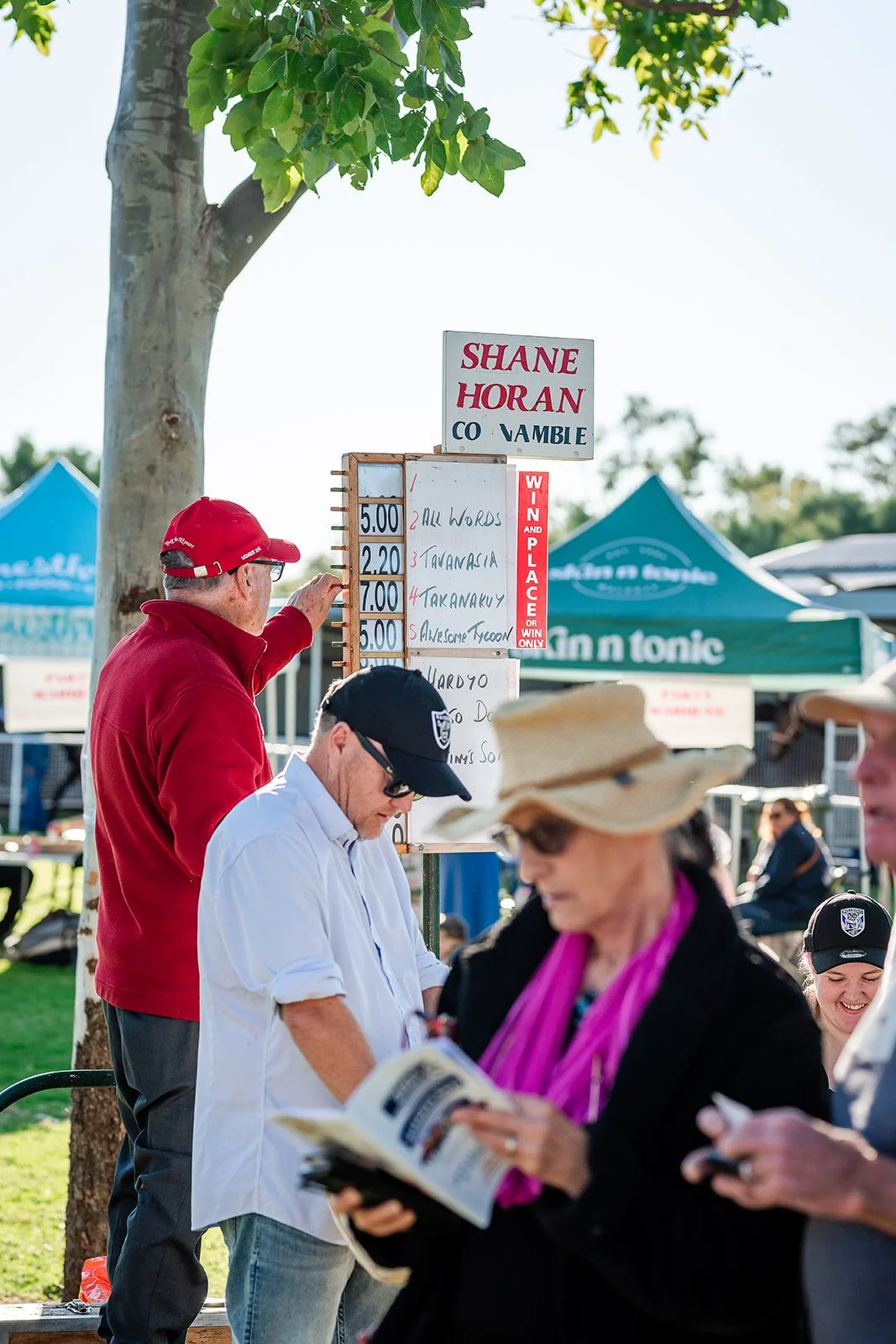 People at the Walgett Races, with a sign showing betting odds for a horse race, tents in the background, and individuals reading or looking at their phones.