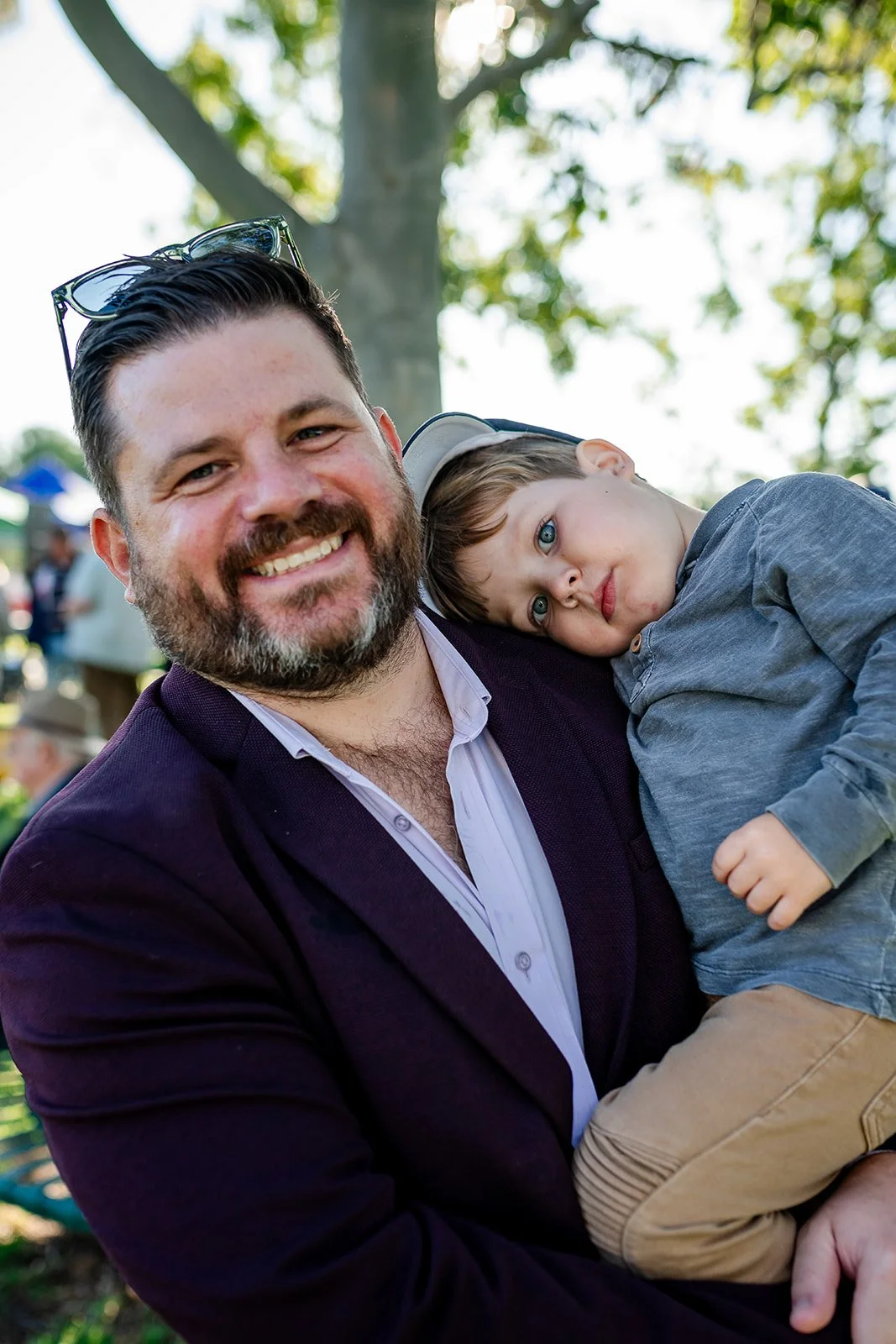 A smiling man with dark hair and beard holding a young boy with blue eyes, outdoors with trees and sunlight in the background. family fun at the Walgett Races.