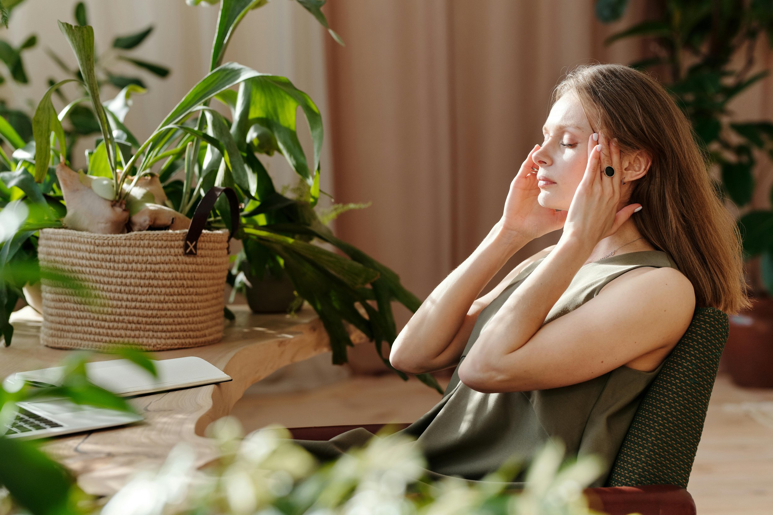 A woman with a headache or migraine sits at a desk with her eyes closed, gently pressing her hands to her head, in a room with lush green plants.