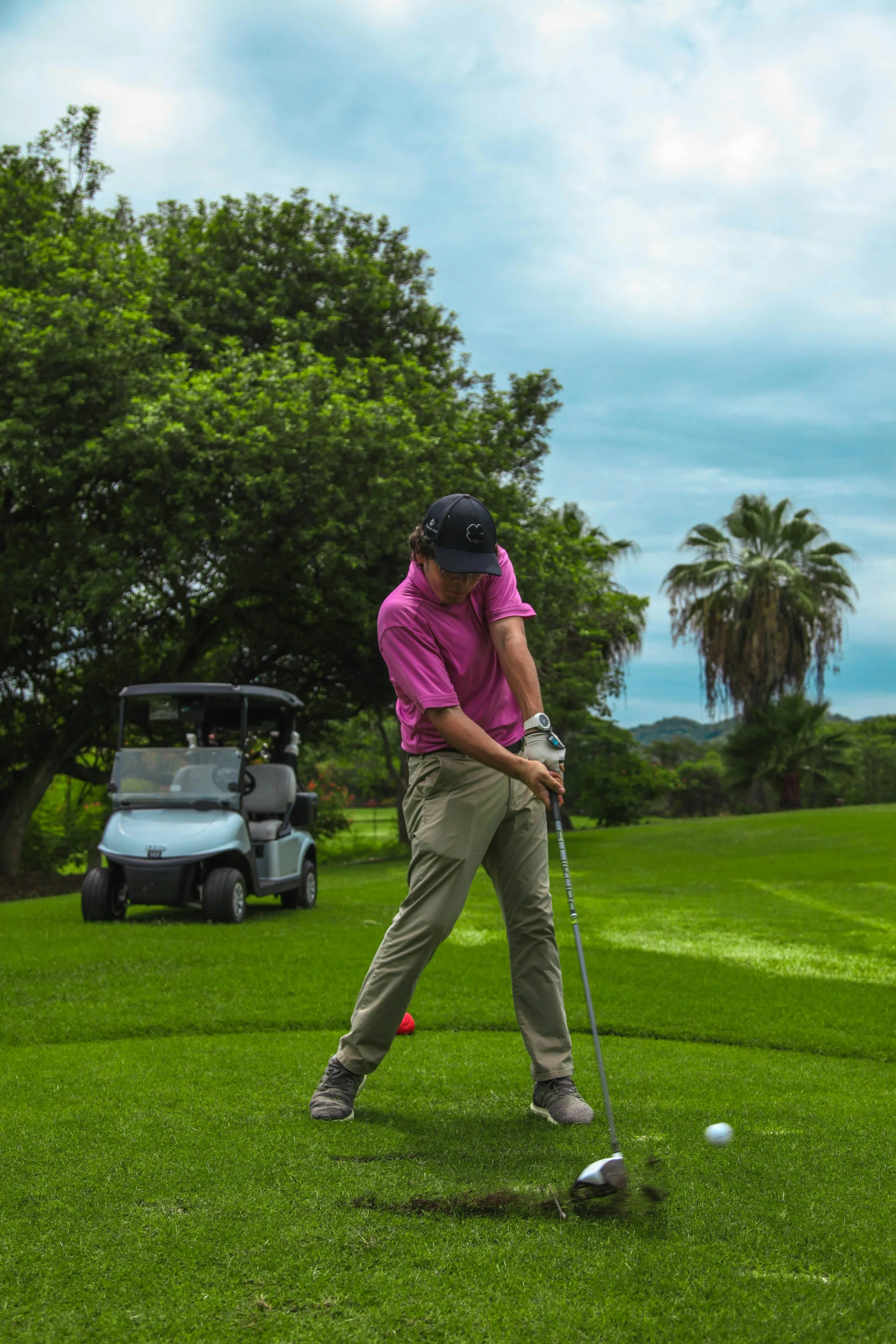 A man wearing a pink shirt, beige pants, and a black cap plays golf on a lush green course. A golf cart is parked behind him, and there are large trees and a cloudy sky in the background.