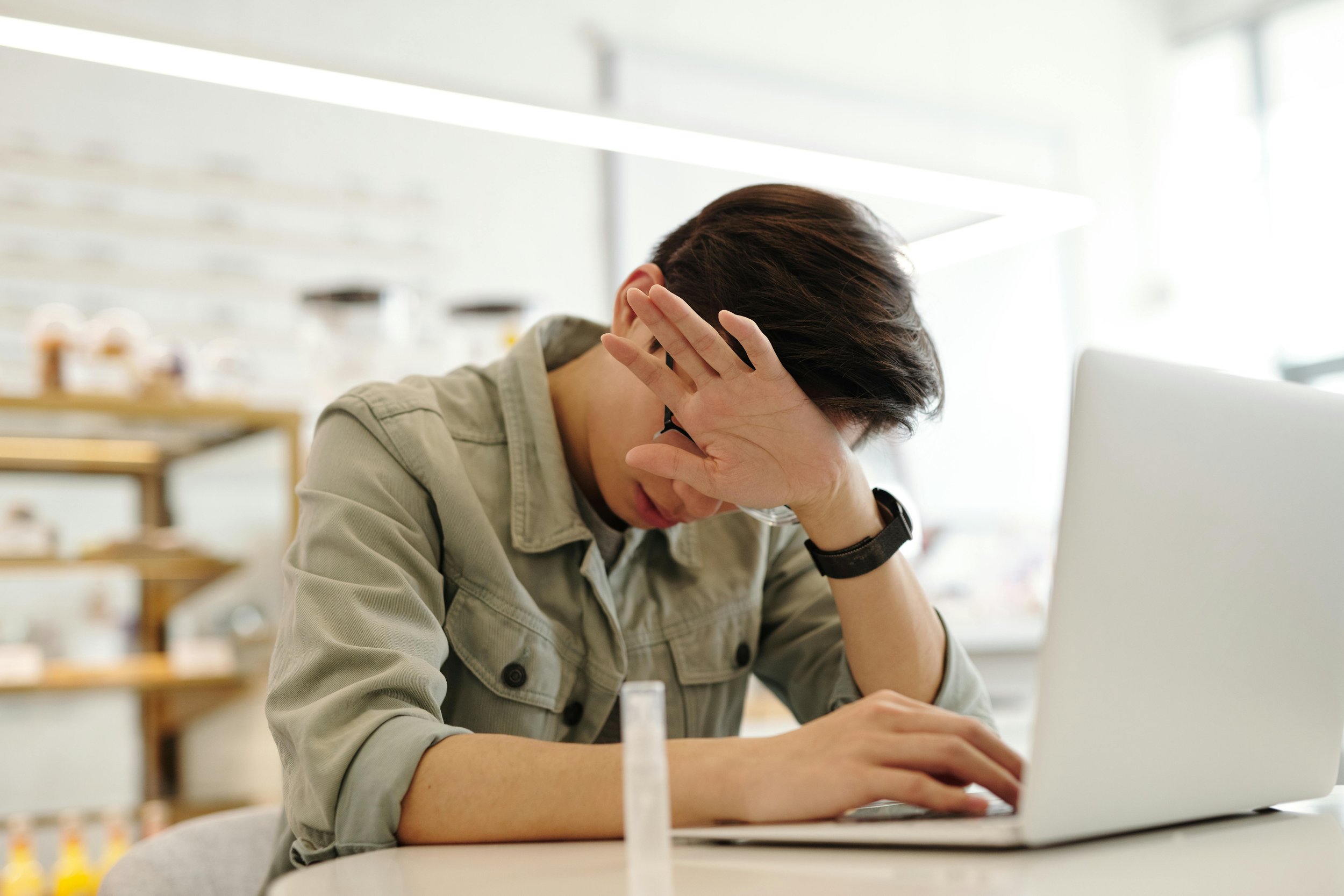 A person sitting at a desk with a laptop, resting their head in their hand, appearing stressed or tired in a bright room.