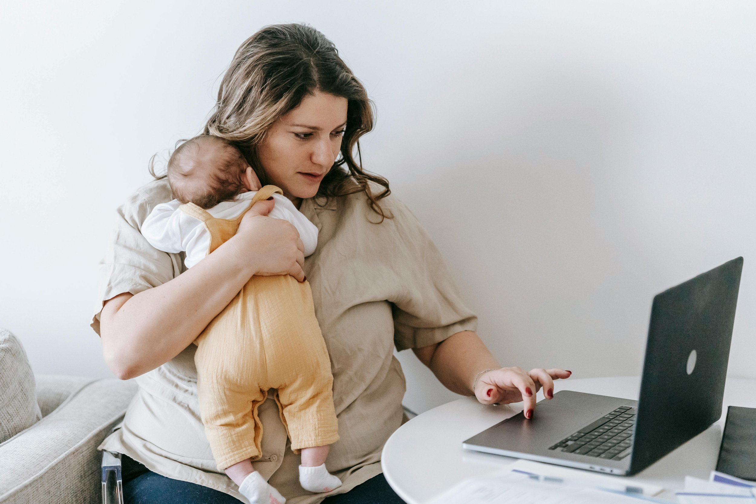 Woman with a baby on her shoulder using a laptop at a table.