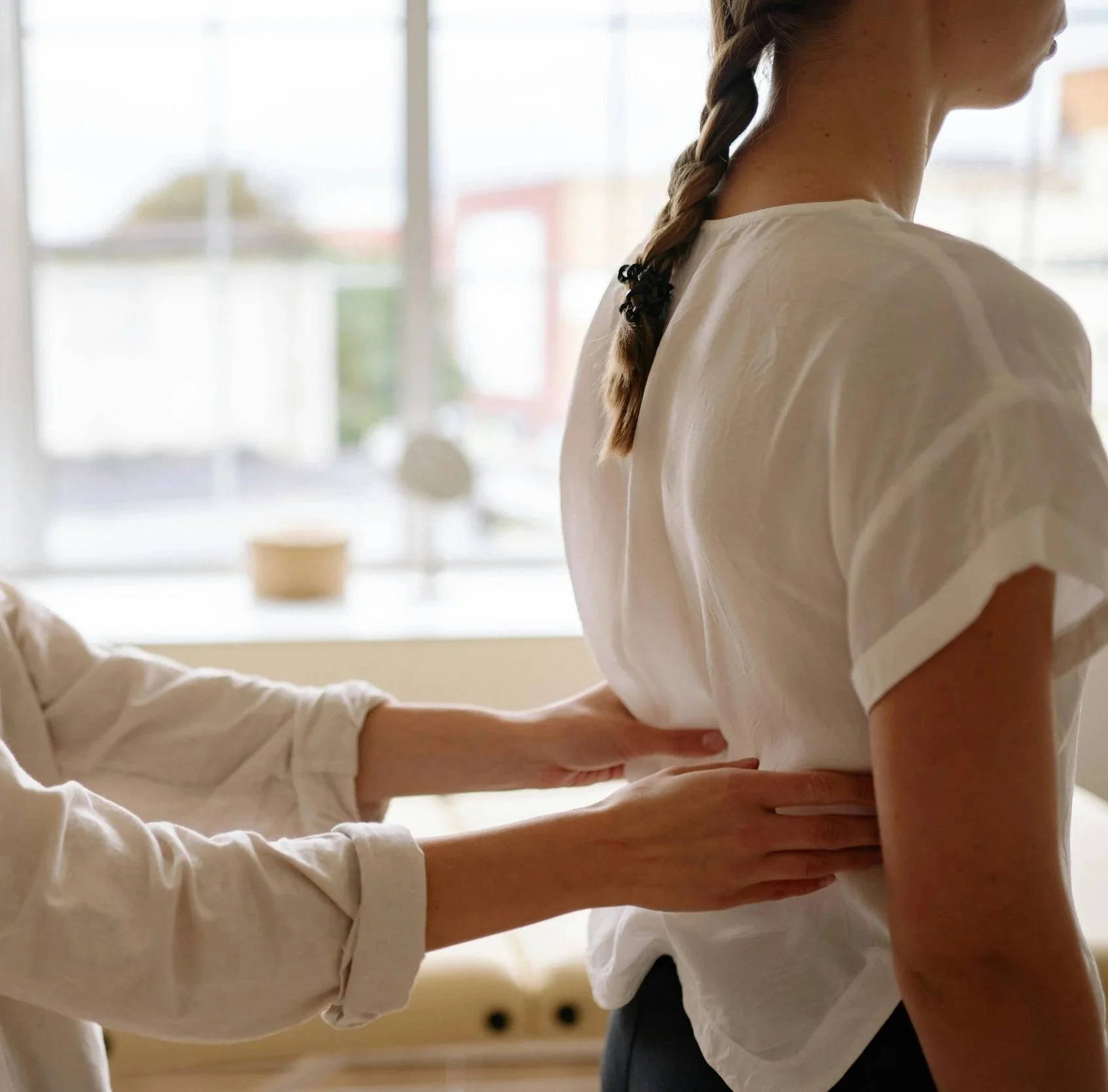 A healthcare professional performs a physical examination or assessment on a woman by placing her hands on the woman's mid-back in a brightly lit room with large windows.