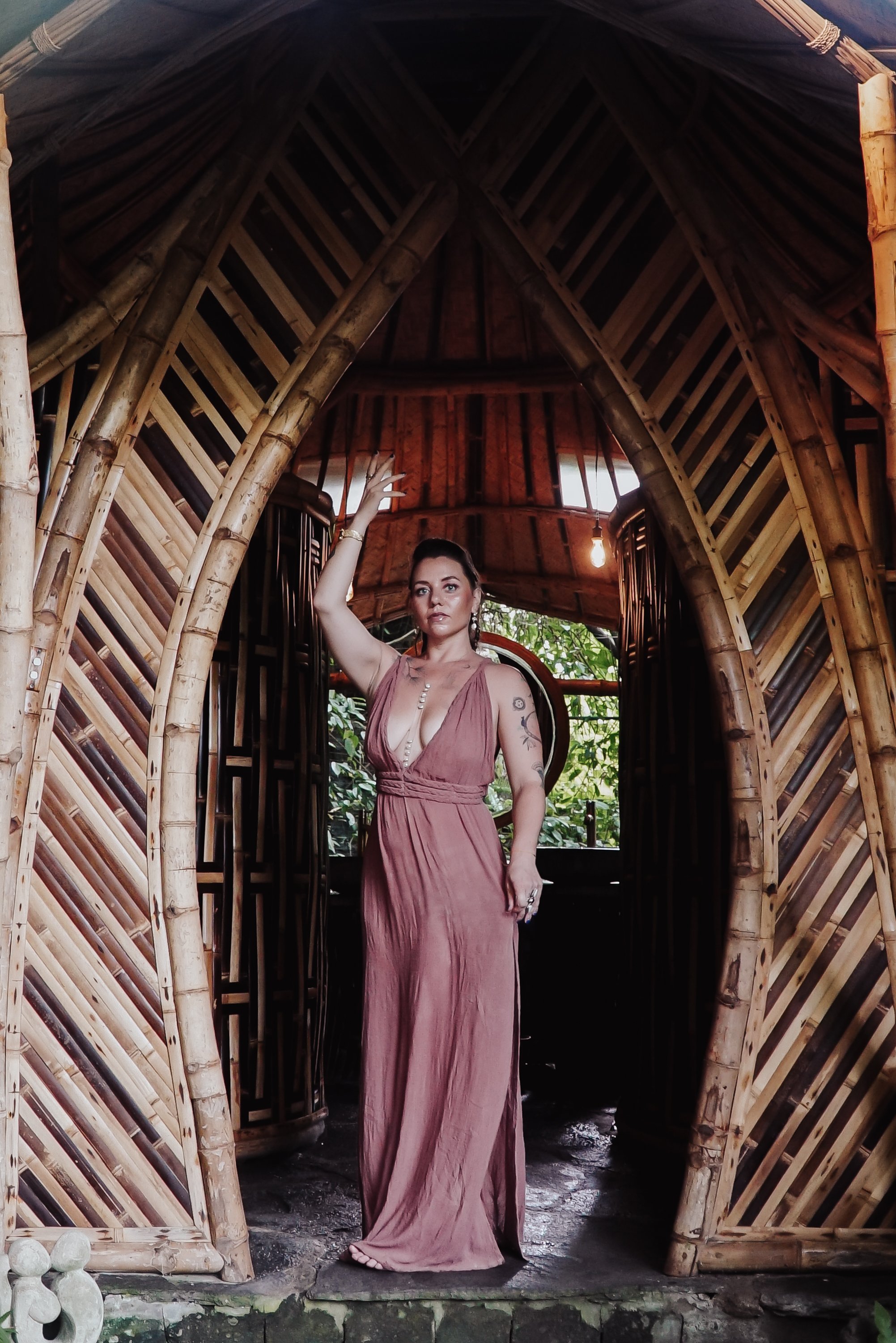 Woman in a flowing pink dress standing in a bamboo gazebo, with her right arm raised and her left hand resting on her hip. The structure has a curved bamboo archway and the background is greenery.