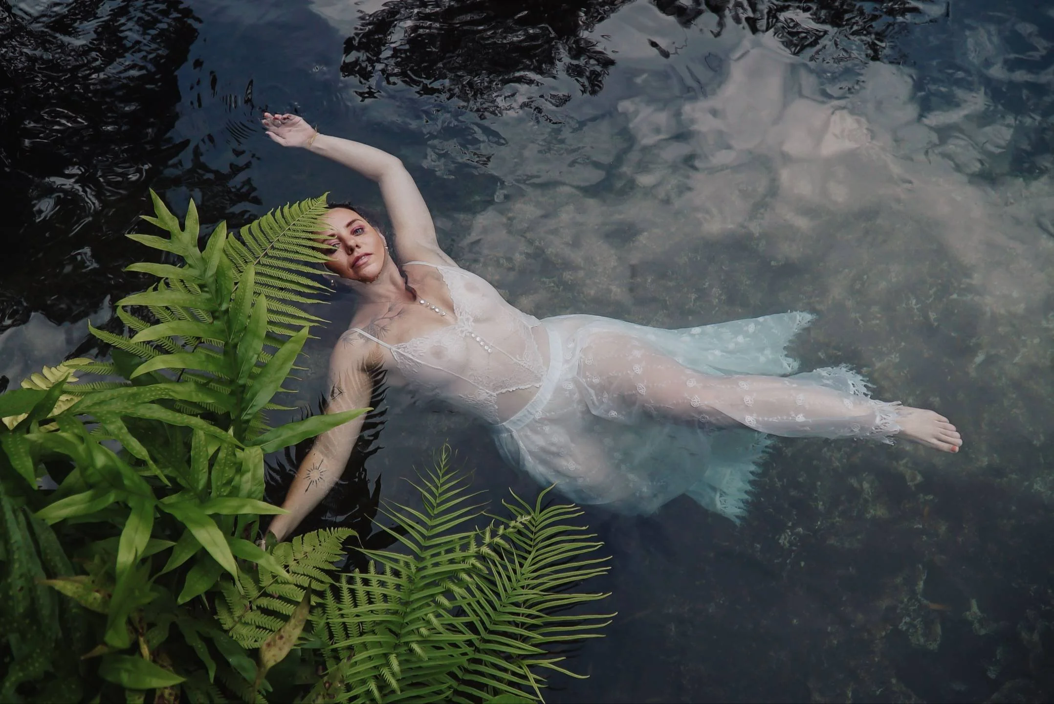 A woman in a white dress and jewelry is floating on her back in a body of water surrounded by green ferns.