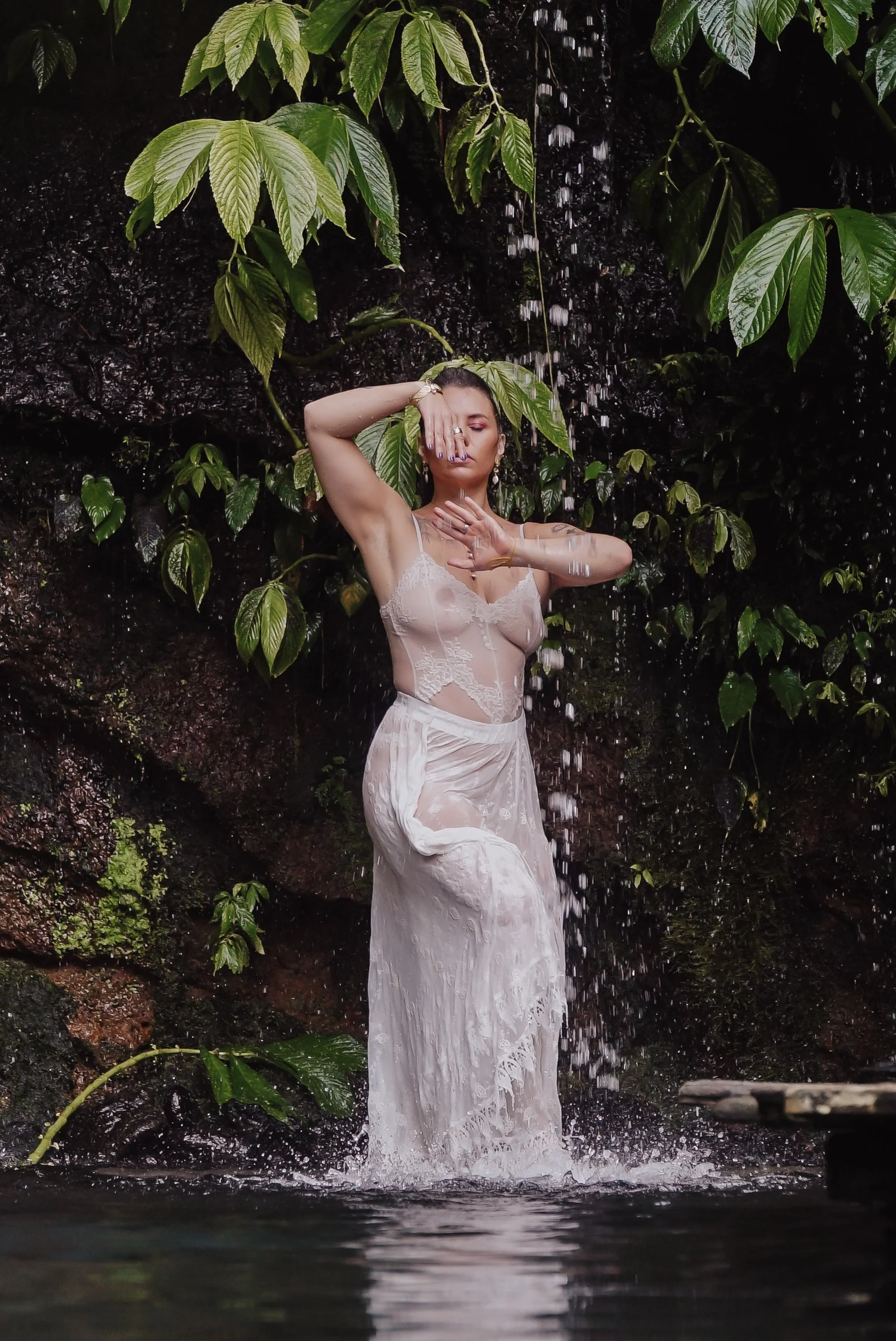 A woman in a sheer, lace dress stands under a waterfall in a lush, green, tropical environment, with water flowing over her head and body.