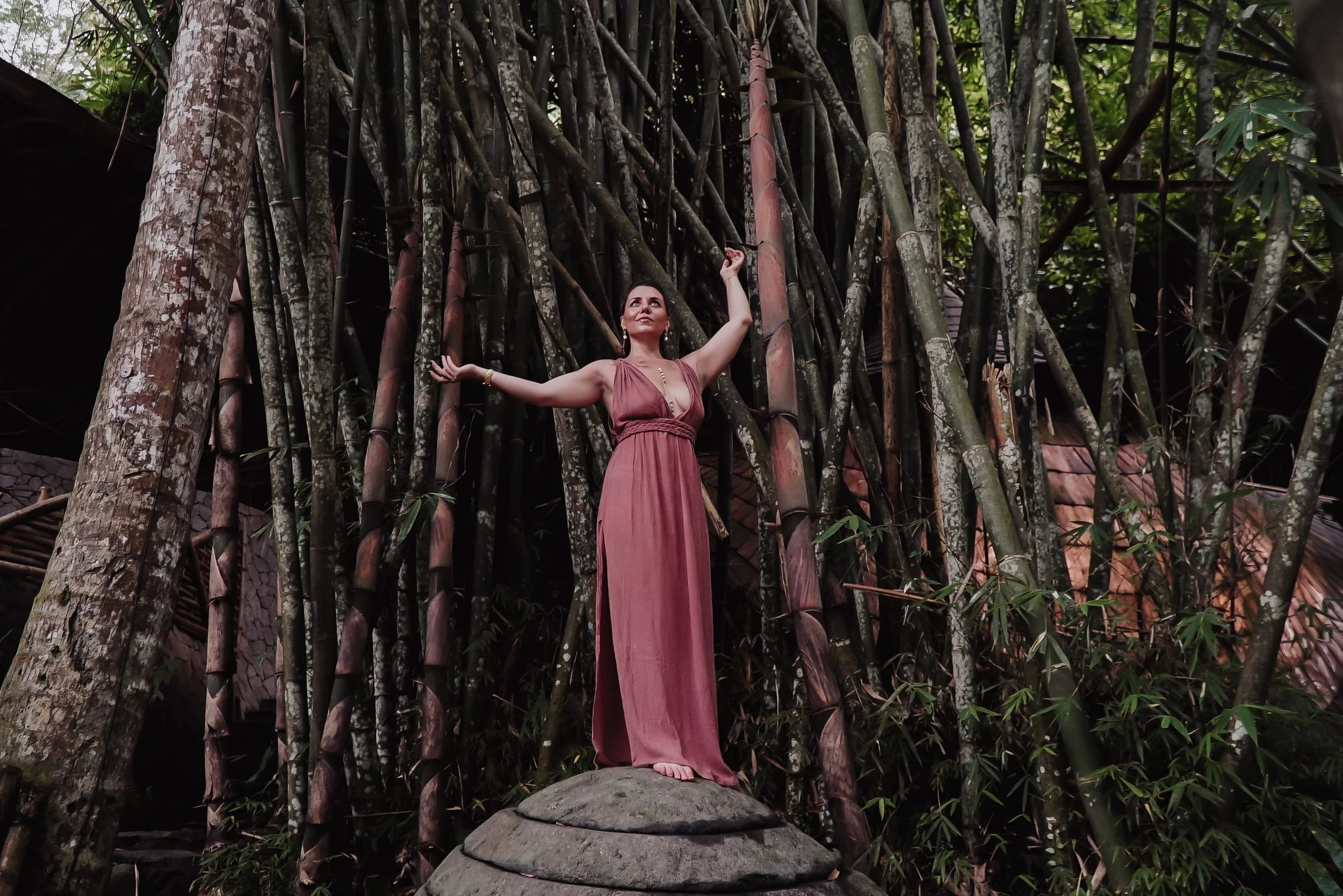 A woman with dark hair and a dusty pink dress standing barefoot on a rock, arms raised, in front of a large bamboo and tree background in a tropical setting.
