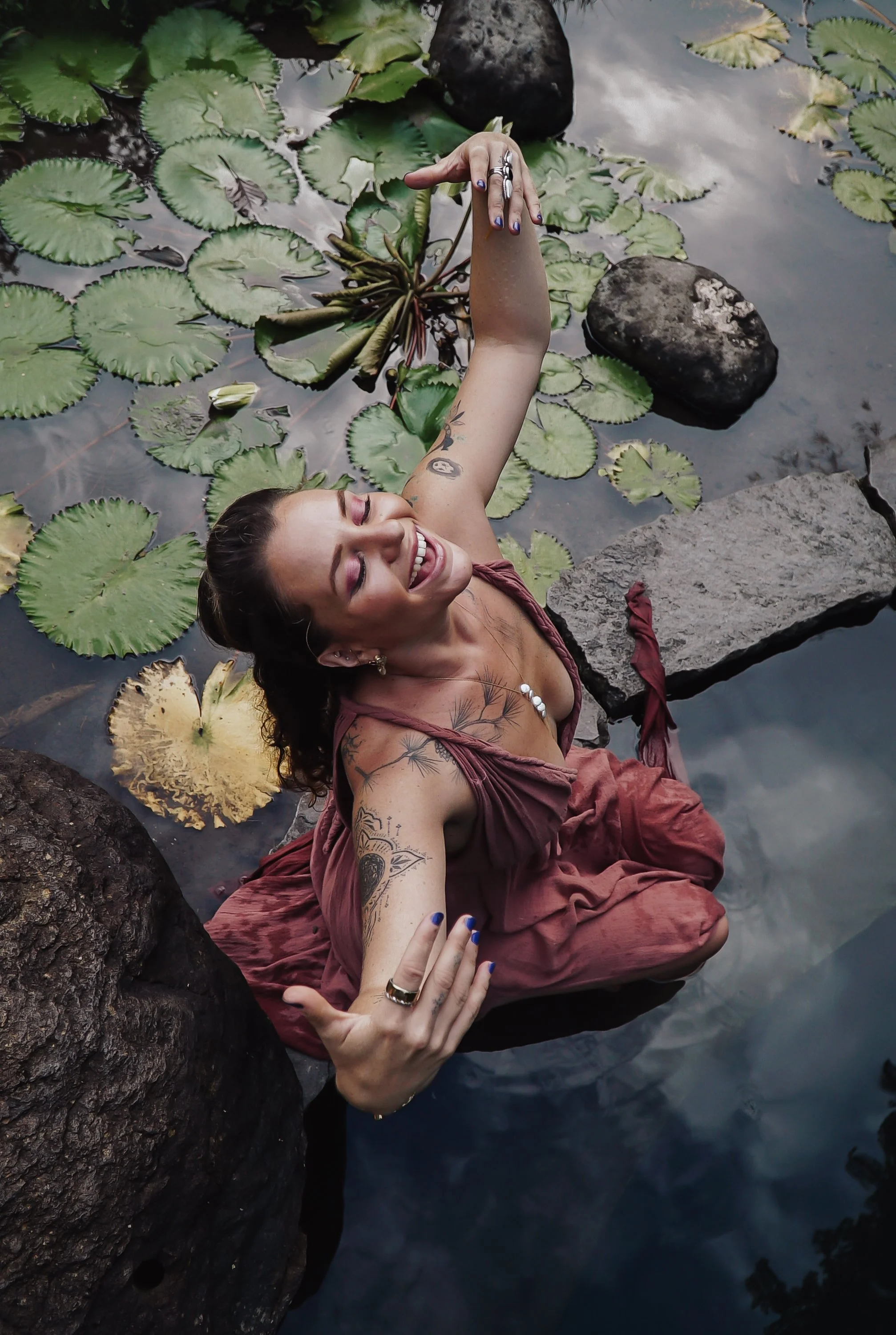 Woman with tattoos smiling, lying among rocks and lily pads in water, taking a selfie with her phone.