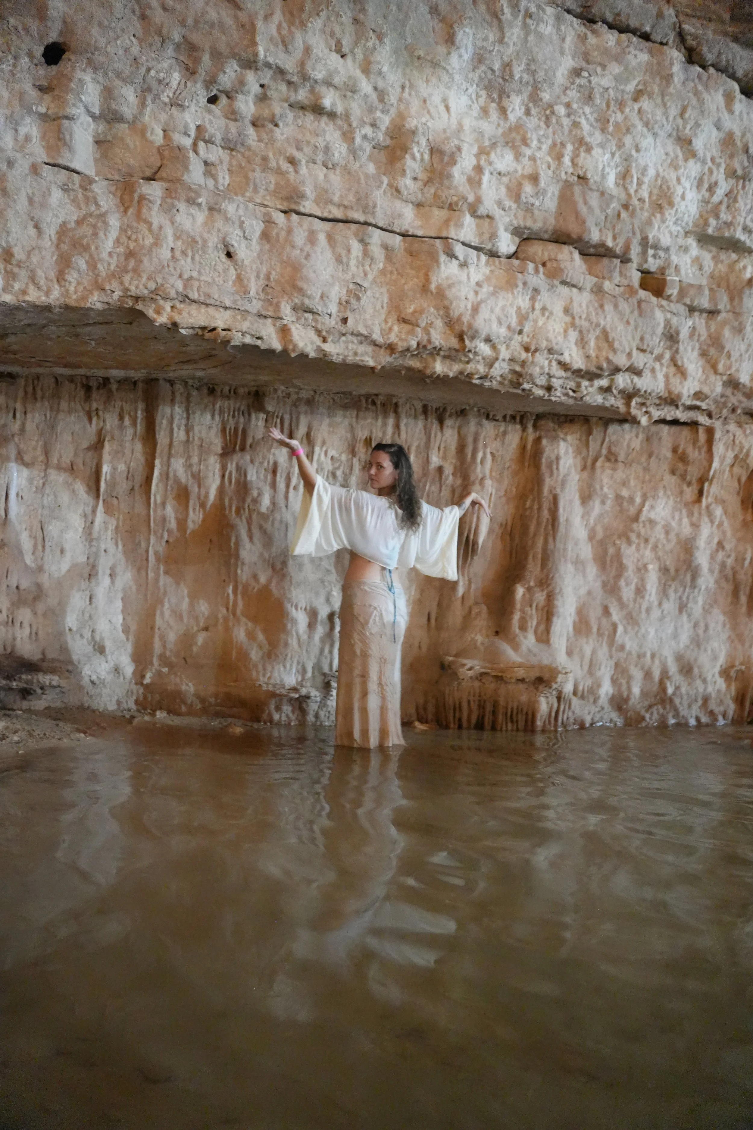 A woman dressed in light-colored clothing standing in a body of water inside a cave with sandstone walls and overhanging rock formations.