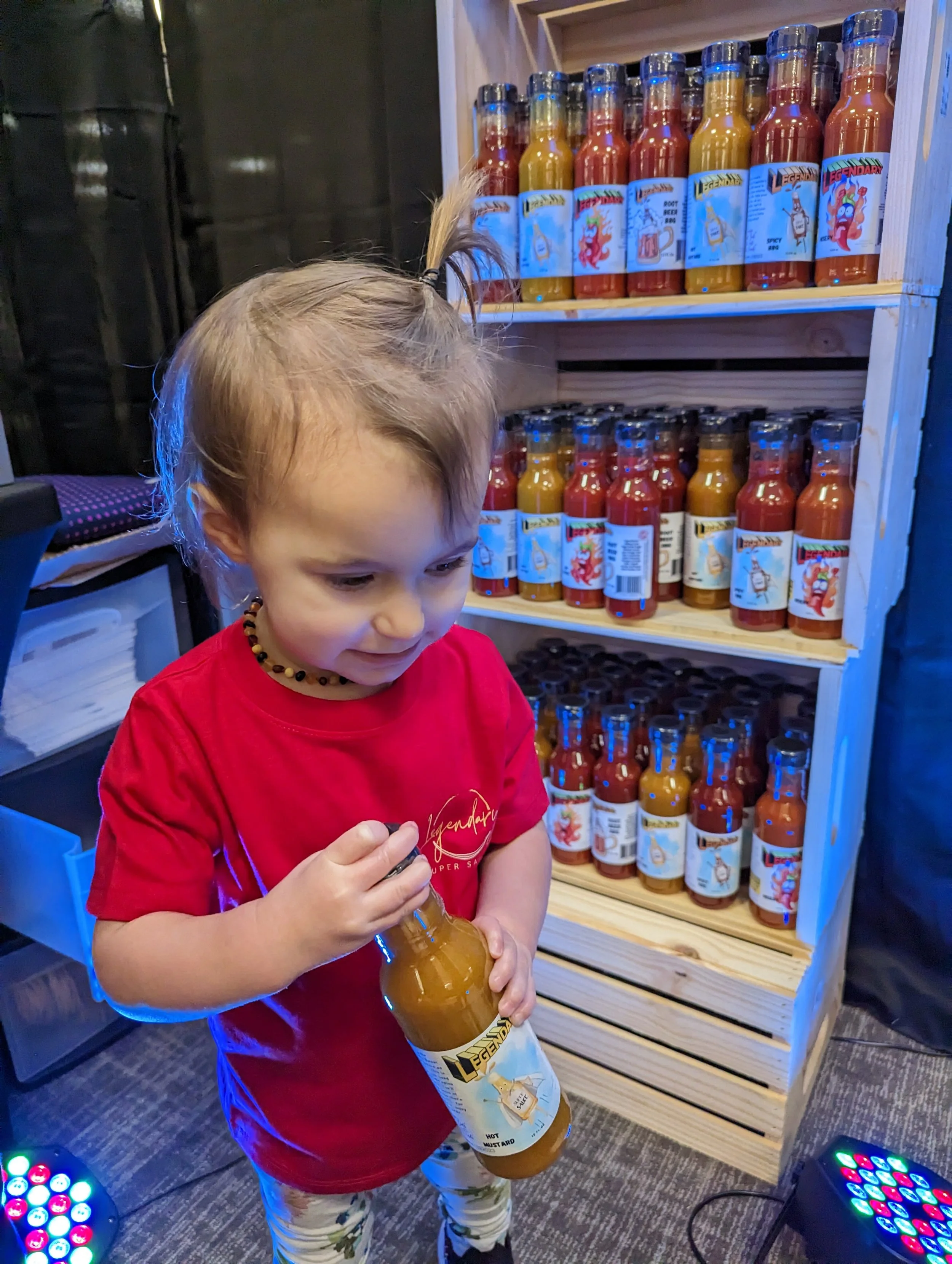 A young girl wearing a red shirt and patterned pants is holding a bottle of Legendary Hot Sauce, examining it, with a shelf of similar bottles behind her. Family Business.