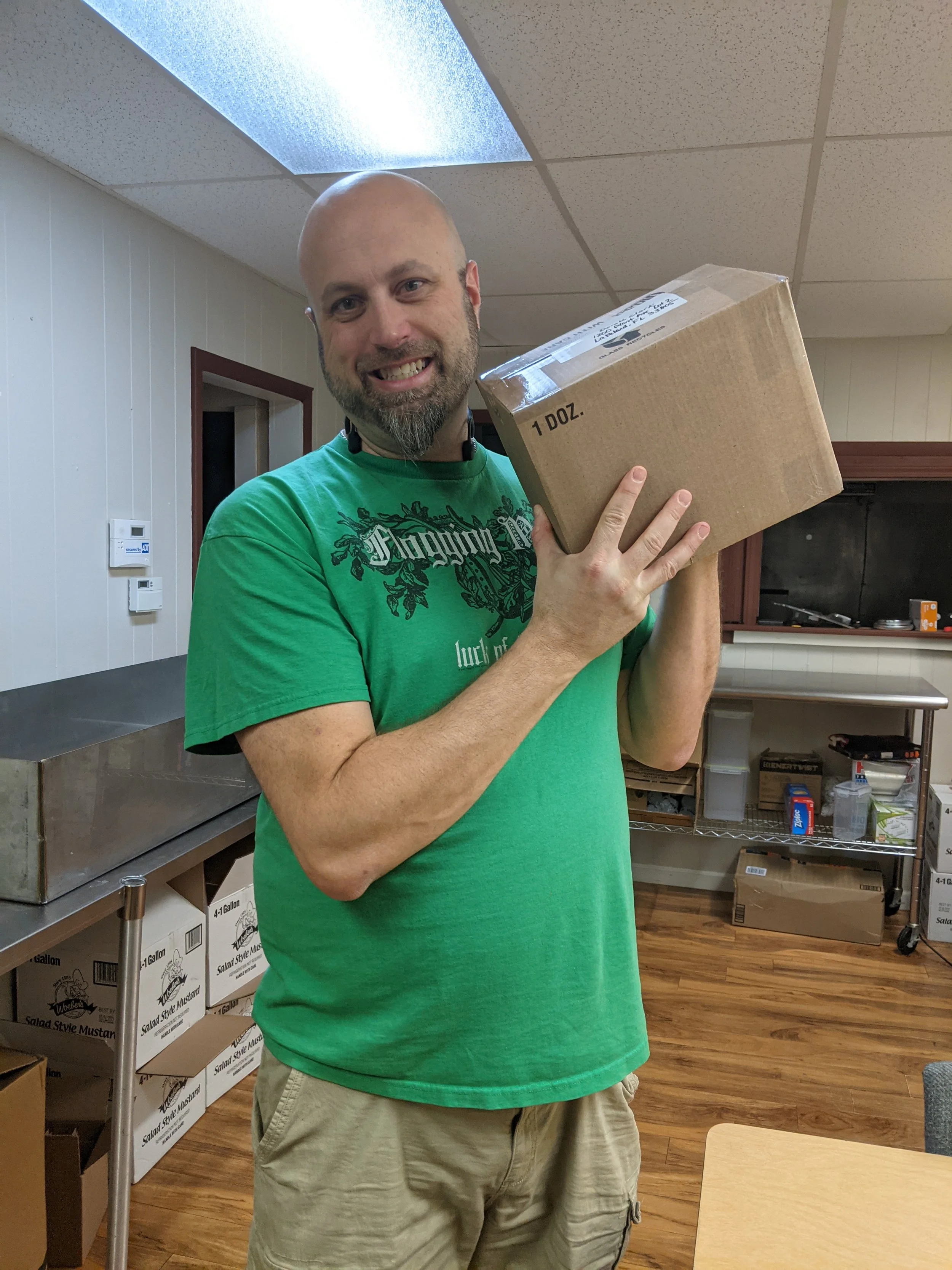 A man with a beard and bald head smiling while holding a cardboard box indoors. He is wearing a green T-shirt and beige shorts, standing in a room with boxes and shelves. Owner of Legendary Super Sauce. Jerry Karns.