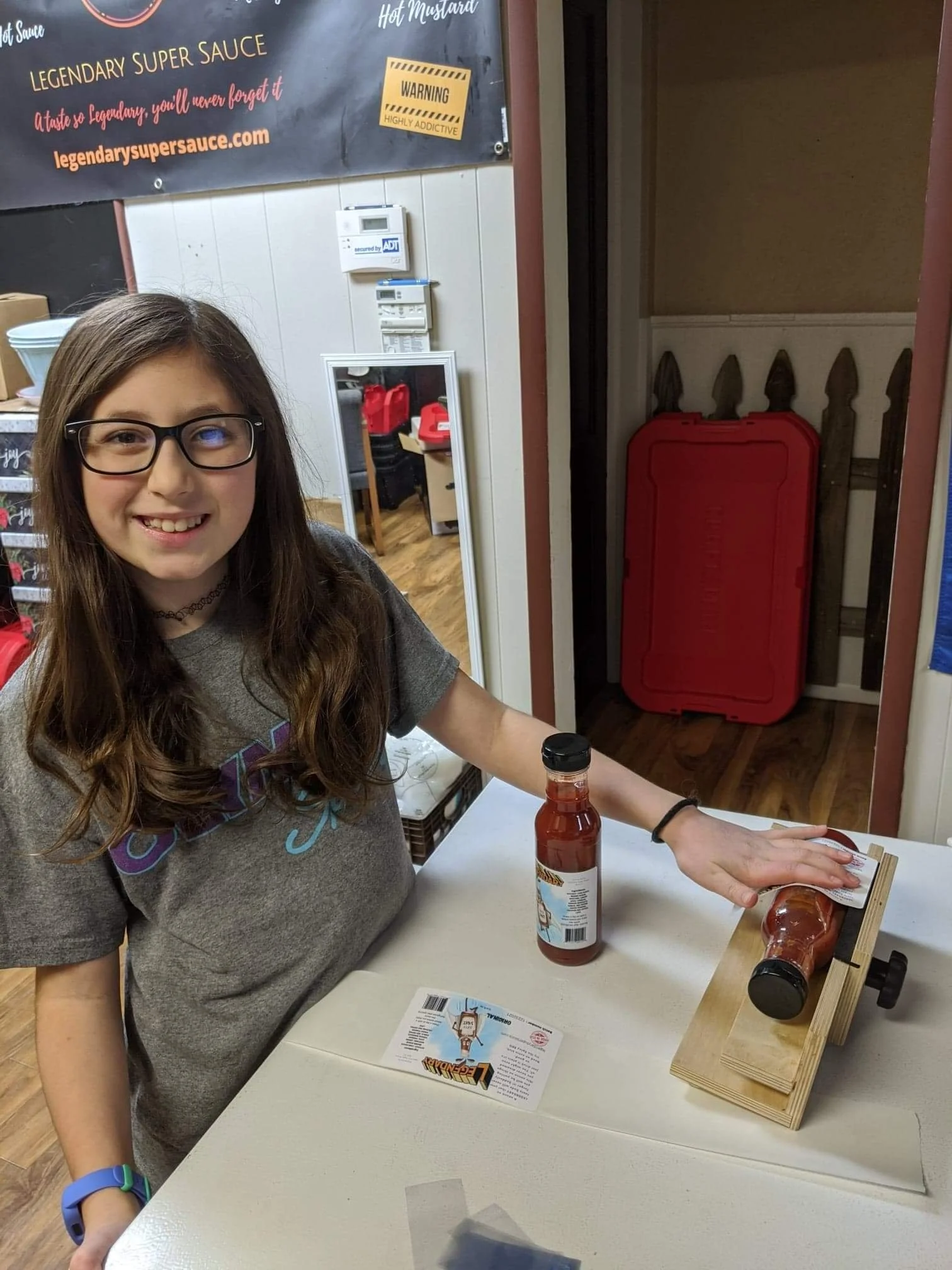 A young girl with long brown hair, glasses, and a gray T-shirt is smiling at the camera while bottling Legendary Super Sauce.  There are additional bottles of sauce in the background waiting to be labeled.