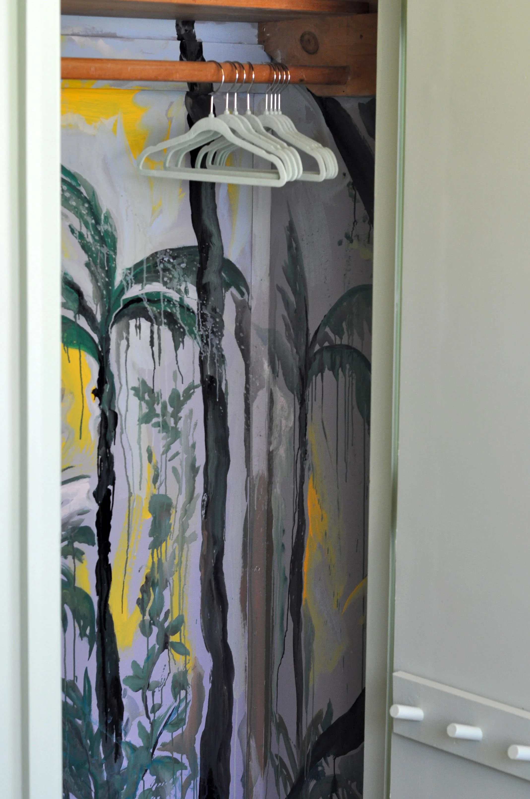 Inside a closet with a painted mural of a jungle scene featuring trees and leaves in green, yellow, and black colors. White hangers are hanging on a wooden rod.