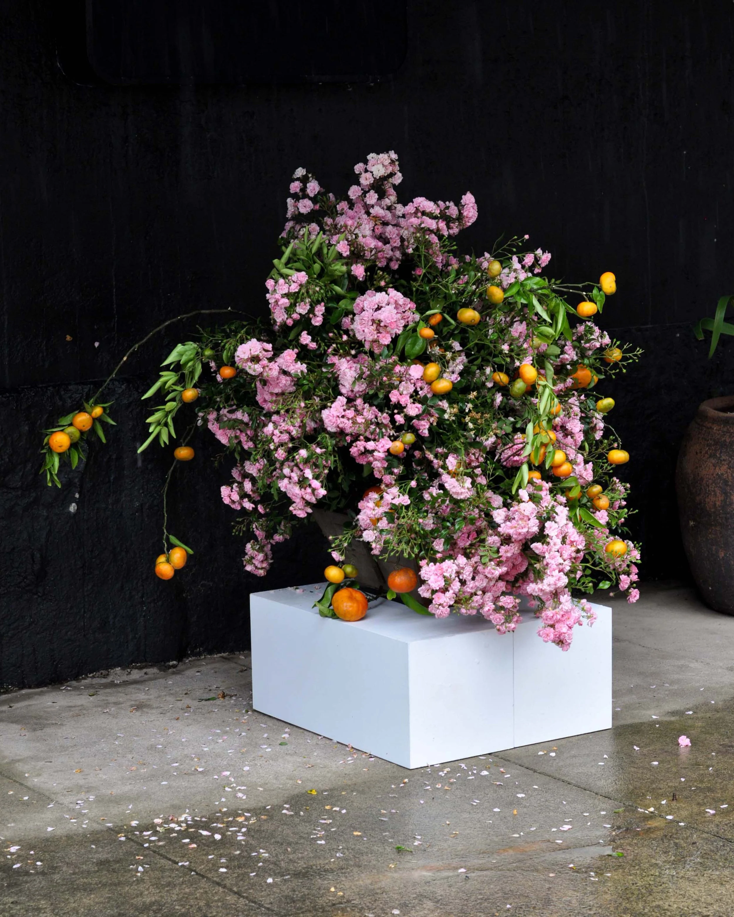 A large floral arrangement with pink flowers and yellow-orange fruits on a white rectangular pedestal against a black wall, with fallen flower petals on the ground.