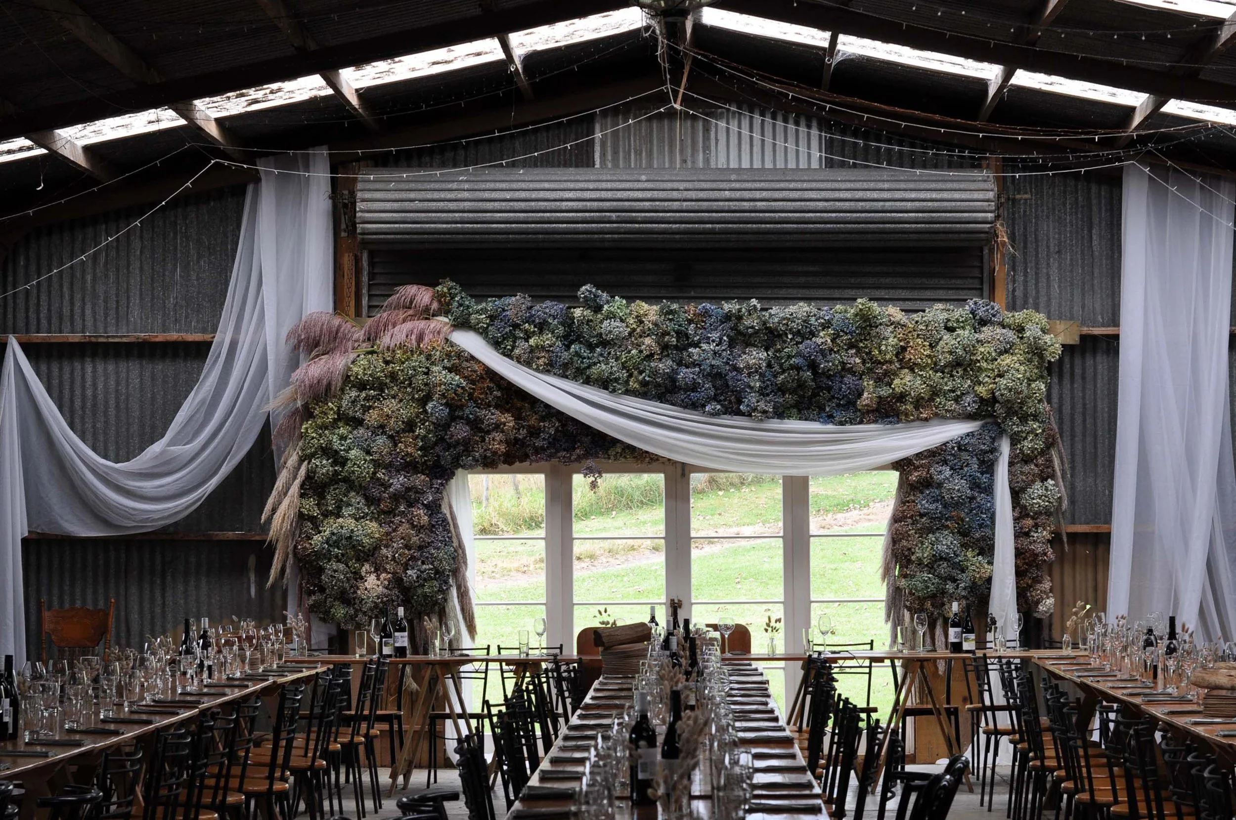 Inside a rustic barn decorated for a wedding, with a high ceiling, string lights, white curtains, and a floral archway over a window, with tables set for a celebration.
