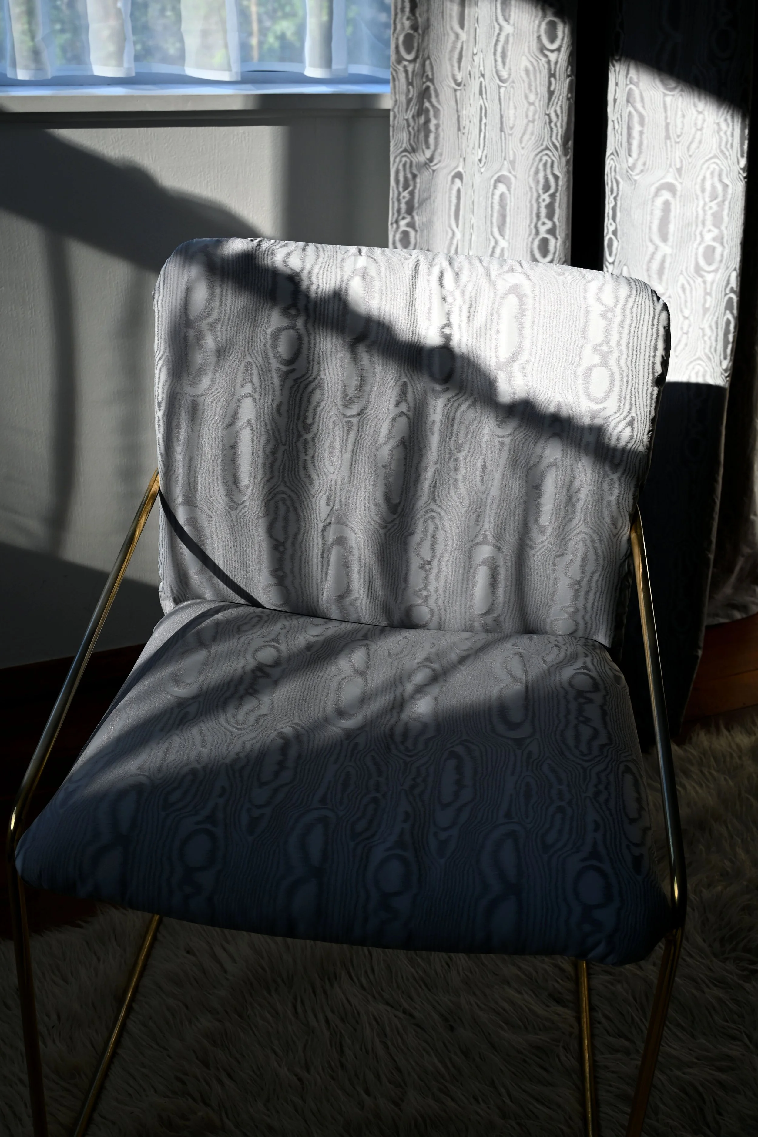 A chair with a patterned fabric covering and gold-colored legs, placed near a window with sheer curtains. Sunlight casts shadows on the chair and walls, and part of a shaggy white rug is visible on the floor.