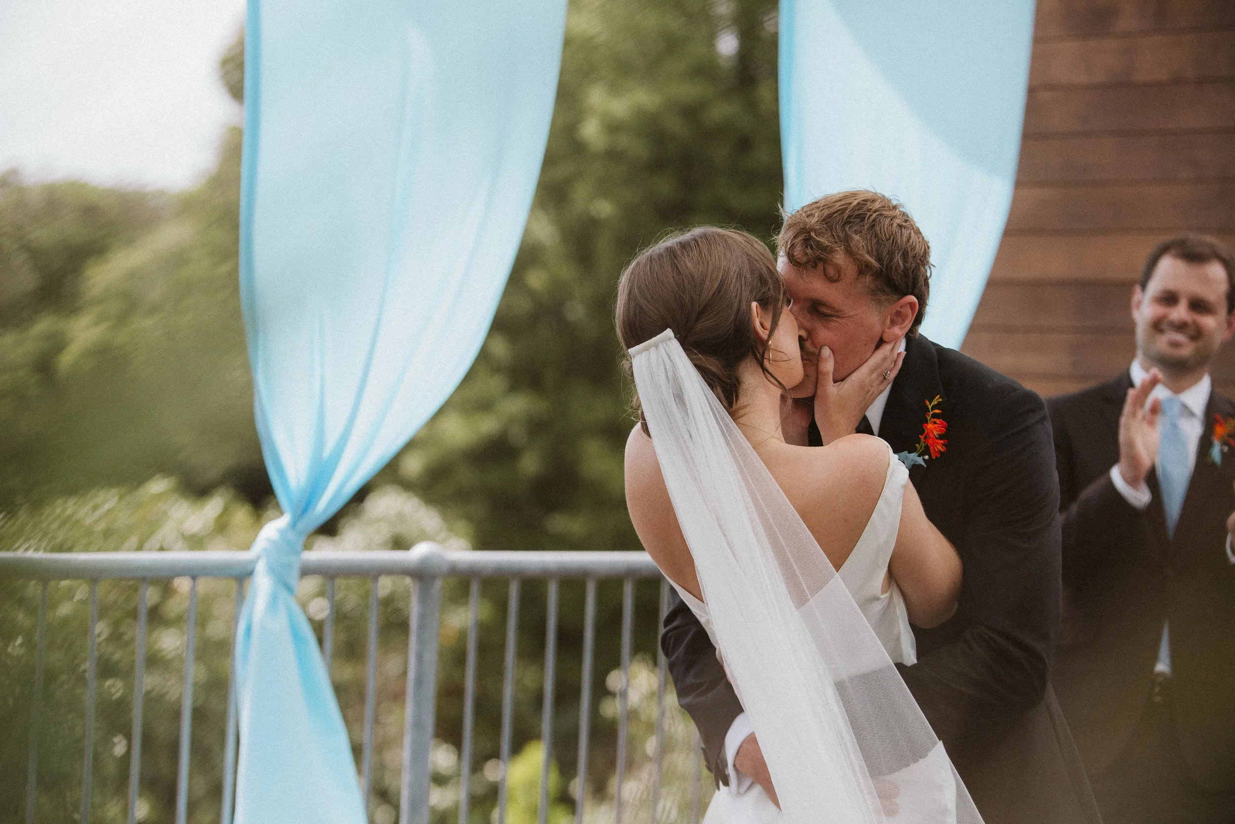 A bride and groom share a kiss during their outdoor wedding ceremony, with a officiant clapping in the background.