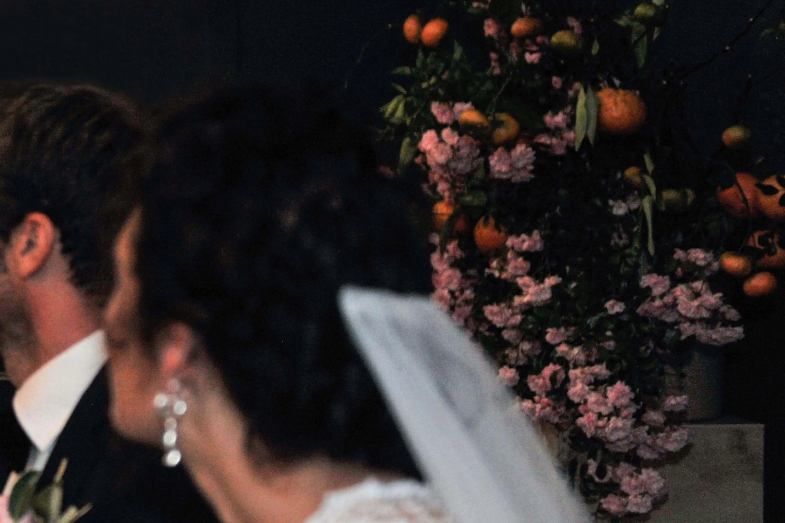 A woman wearing a white dress, black jacket, pearl necklace, and earrings, standing in front of a floral arrangement with pink and orange flowers.