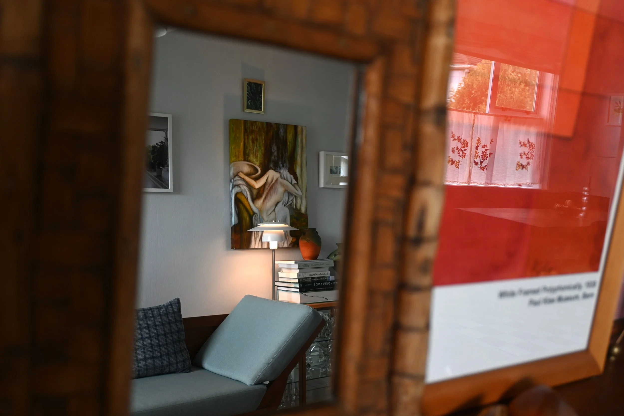 A close-up view through a mirror showing a living room with a painting on the wall, a table lamp, a stack of books, and a window with red curtains.