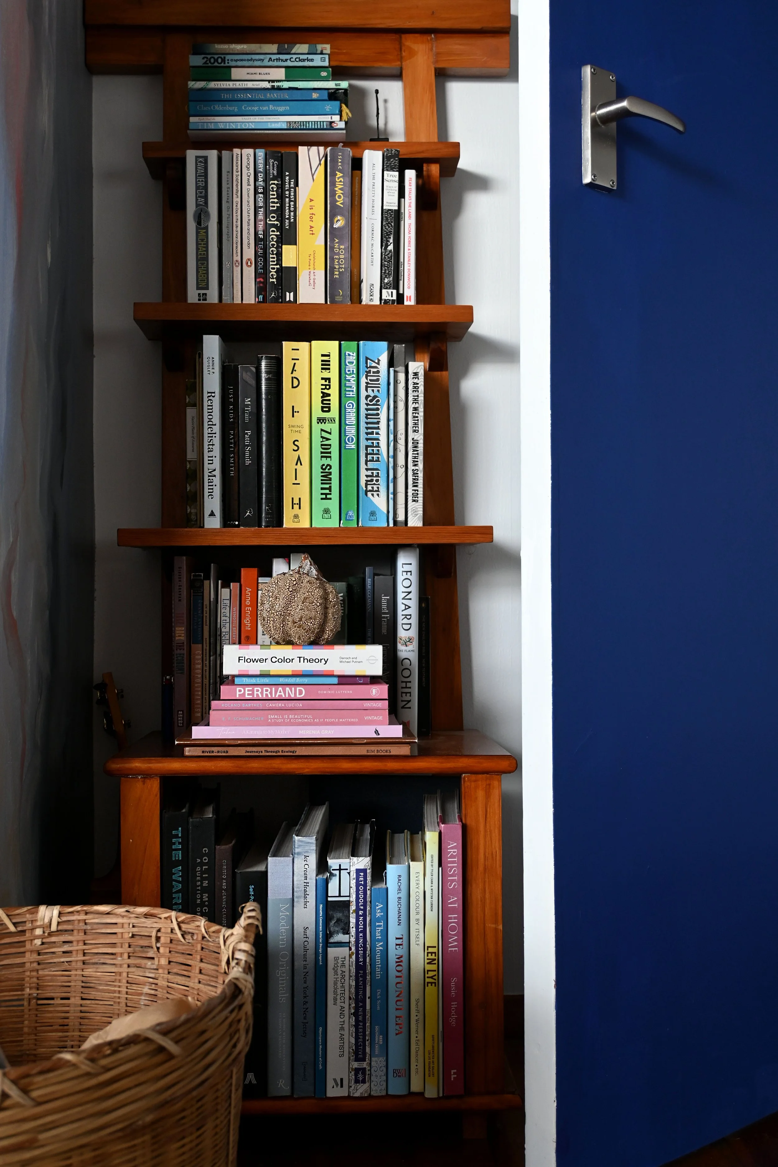 A wooden bookshelf filled with books against a blue and white wall. There is a wicker basket partially in the foreground.