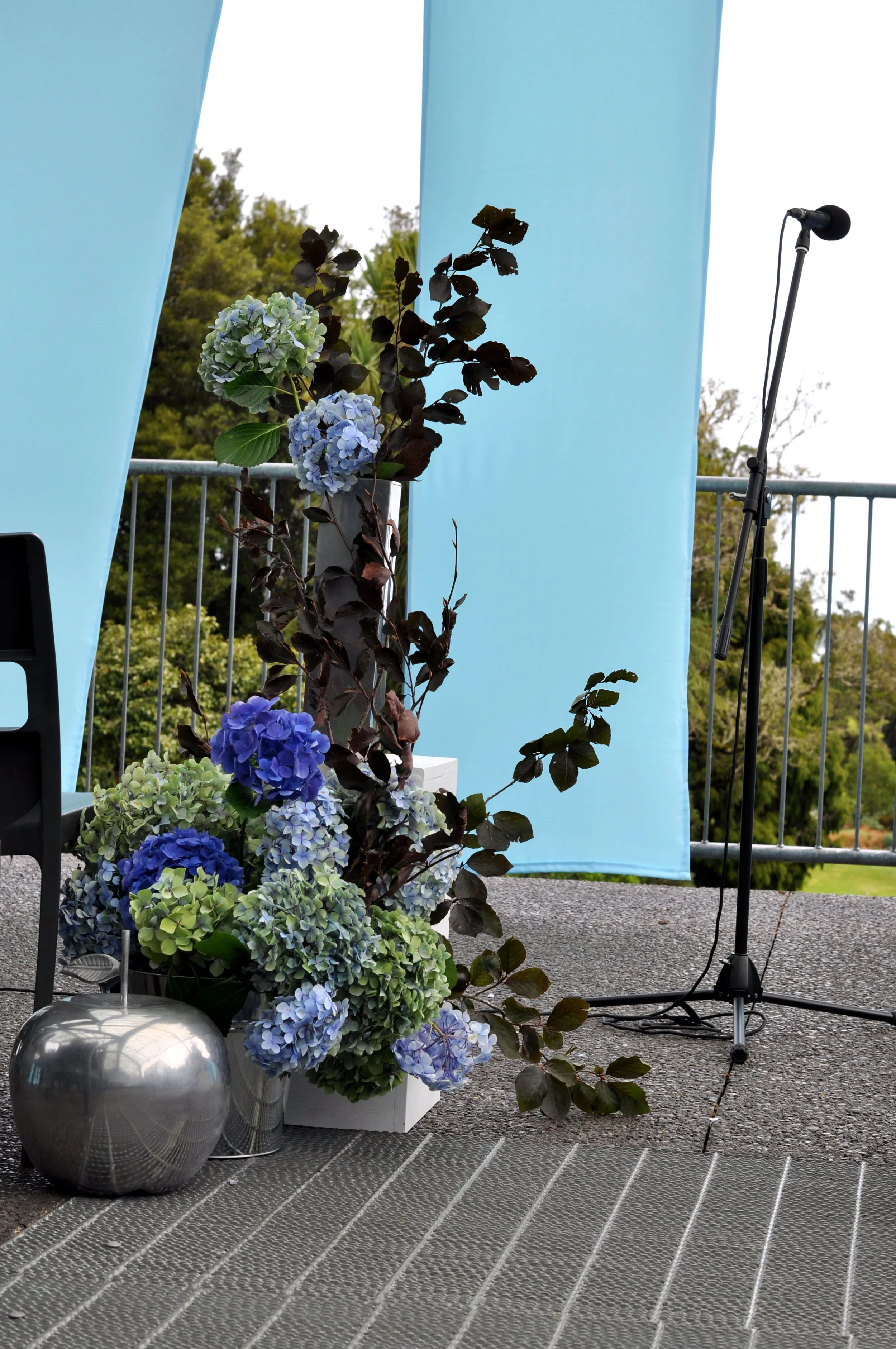 A floral arrangement with blue and green hydrangeas and dark-colored leaves in metal vases on a table, with a light blue backdrop and microphone stand on an outdoor balcony.