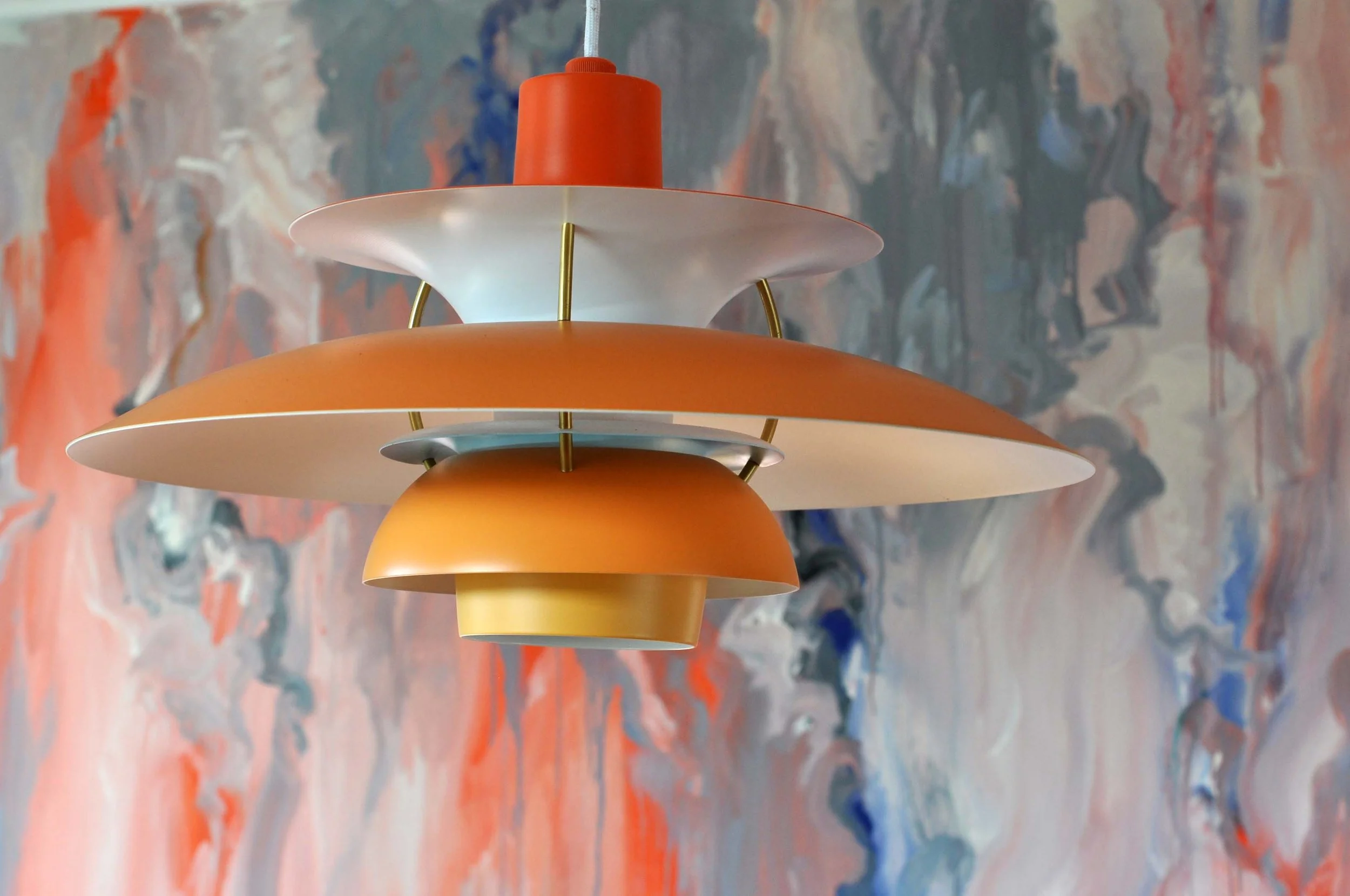 A modern, multi-tiered hanging lamp with an orange, white, and yellow color scheme, hanging in front of a colorful abstract background.
