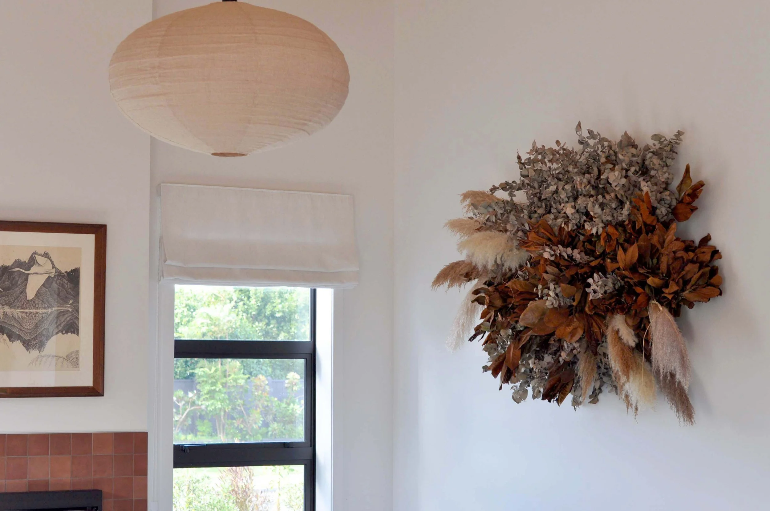 A white wall decorated with a framed black and white landscape photograph, a window with a white curtain, a beige paper lantern ceiling light, and a large dried floral arrangement with brown and muted colors.